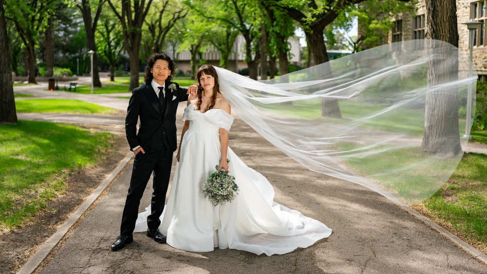 A bride and groom standing together on a park walkway, with the bride's veil flowing in the wind, surrounded by green trees and a city park background.