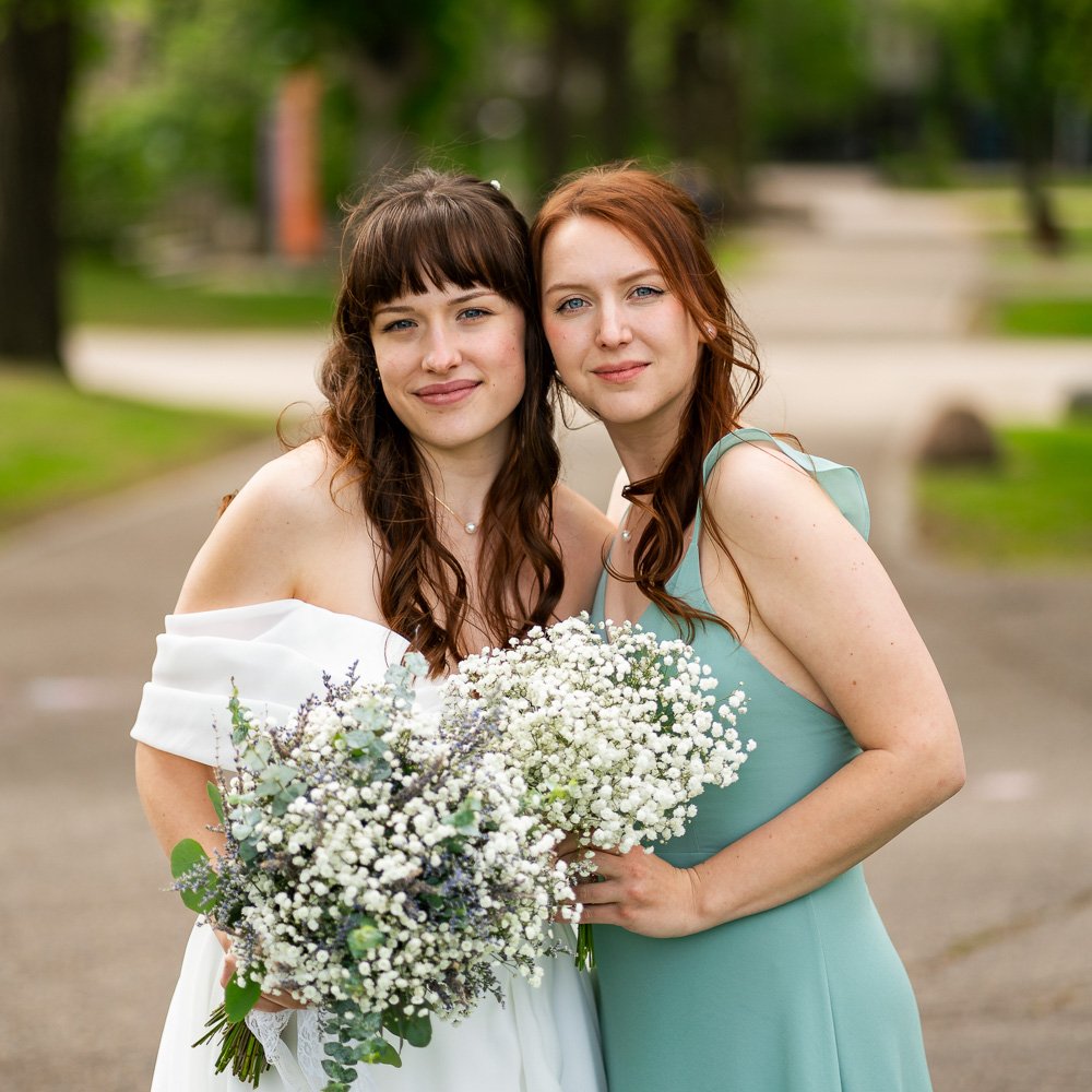 Two women standing close together outdoors, one in a white dress holding a bouquet of white flowers, and the other in a light blue dress, both smiling. Trees and a pathway are in the background.