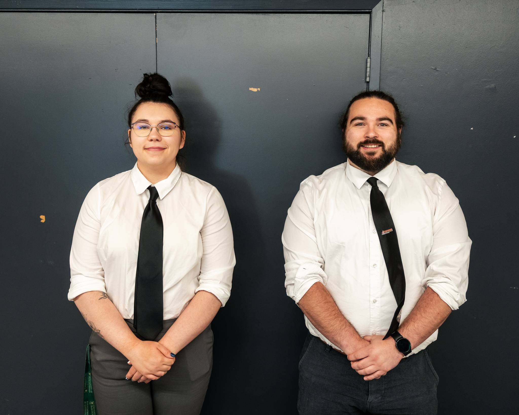 Two people standing in front of a dark gray wall, wearing white shirts and black ties, smiling at the camera.