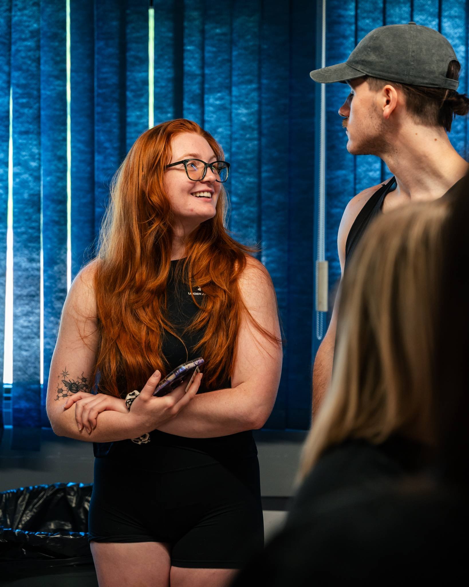 A woman with red hair and glasses smiling while talking to a man wearing a gray cap and black tank top in a room with blue curtains.