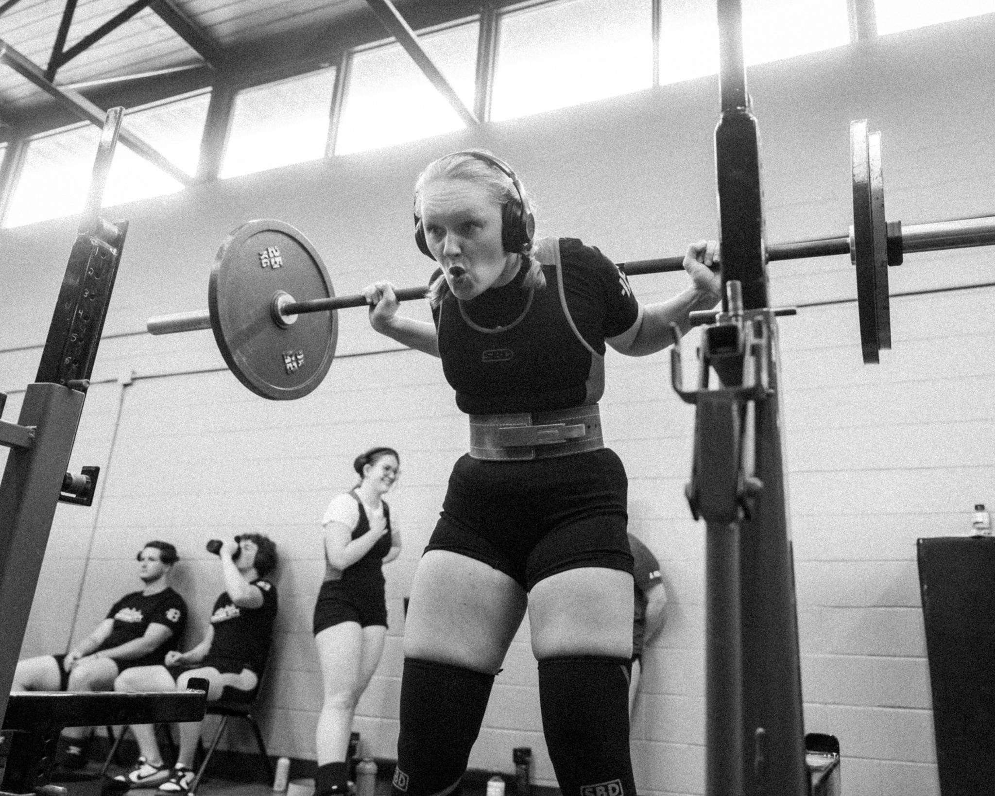 A woman wearing headphones and a lifting belt performs a squat in a gym, with two women sitting and two women standing in the background.