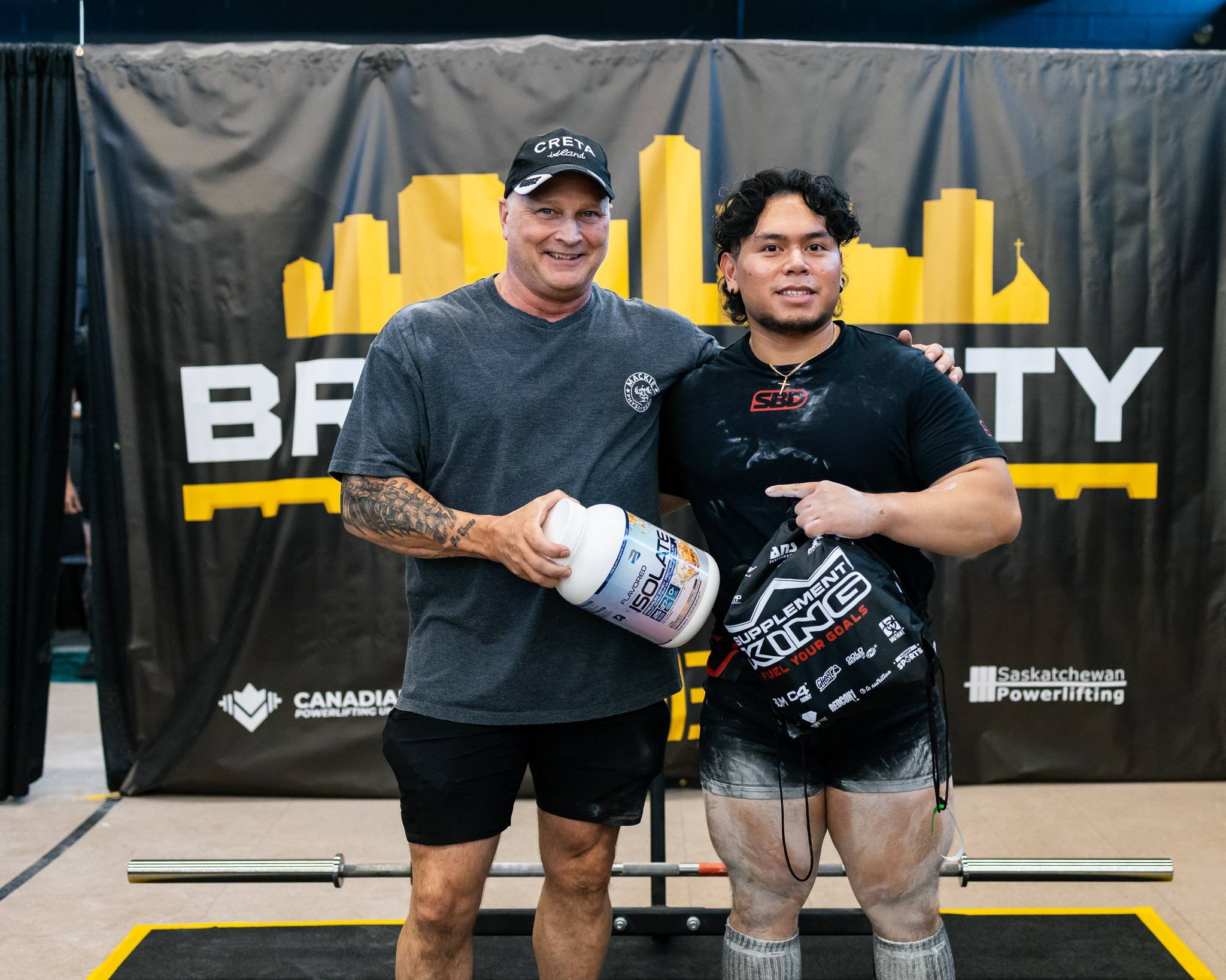 Two men standing together at a powerlifting event; one holding a container of supplements, both smiling, with a black and yellow backdrop displaying the words 'BRY CITY' and logos including 'Canadian Powerlifting' and 'Saskatchewan Powerlifting'.