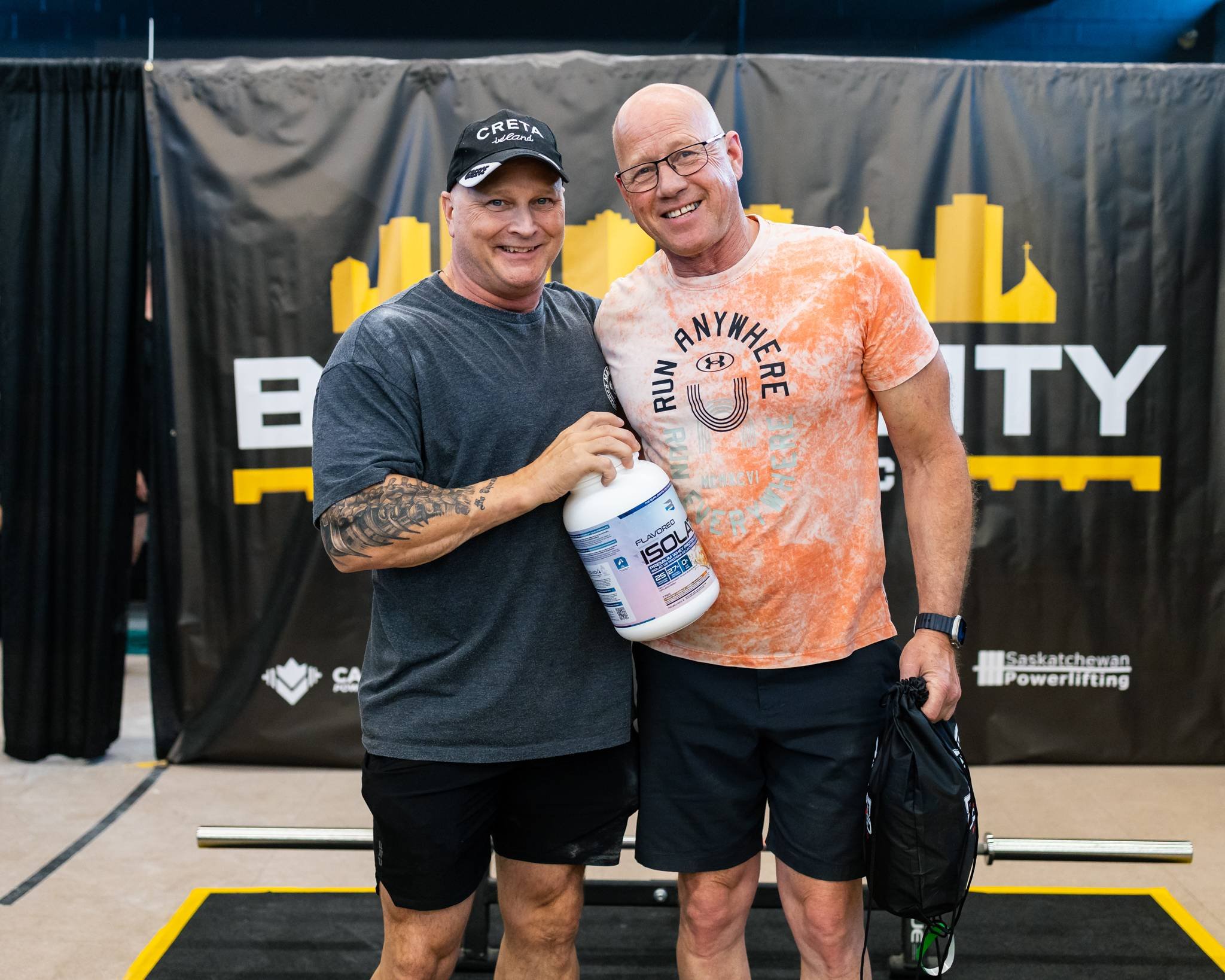Two men smiling and posing together at a fitness event, one holding a container of protein powder, with a black and yellow backdrop behind them.