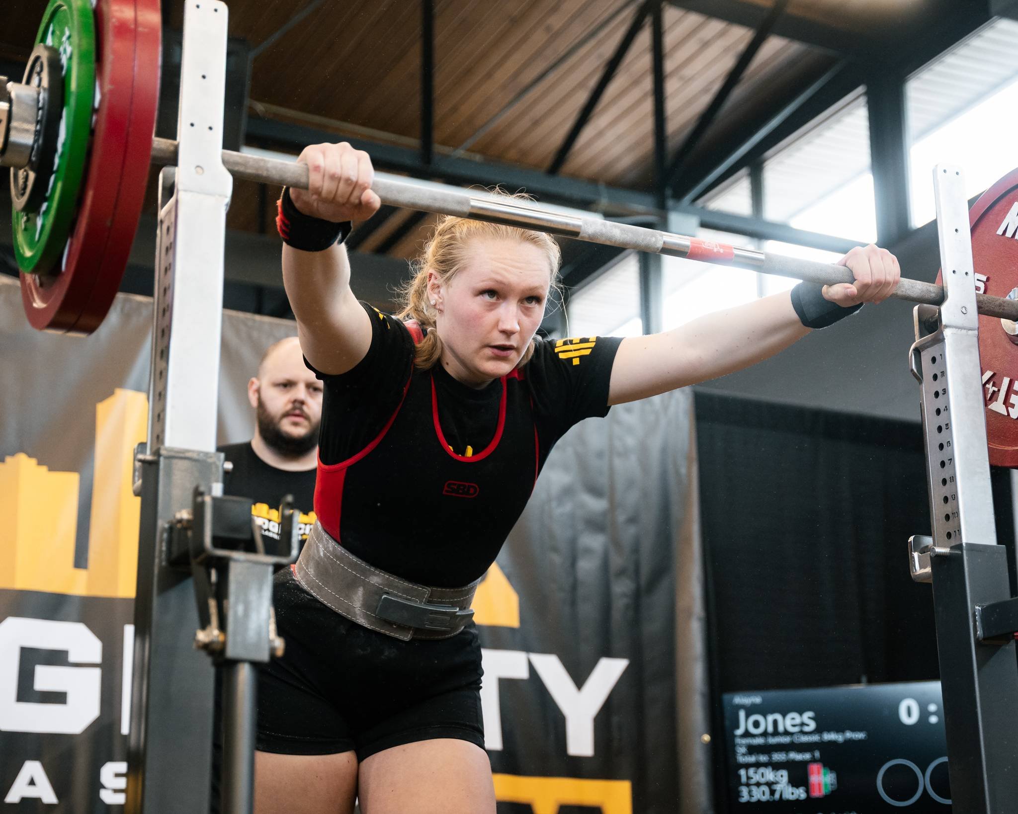 Young woman with blonde hair in a ponytail doing a weightlifting squat at a gym, with a judge or coach standing behind her, wearing a black shirt, black shorts, and a weightlifting belt.