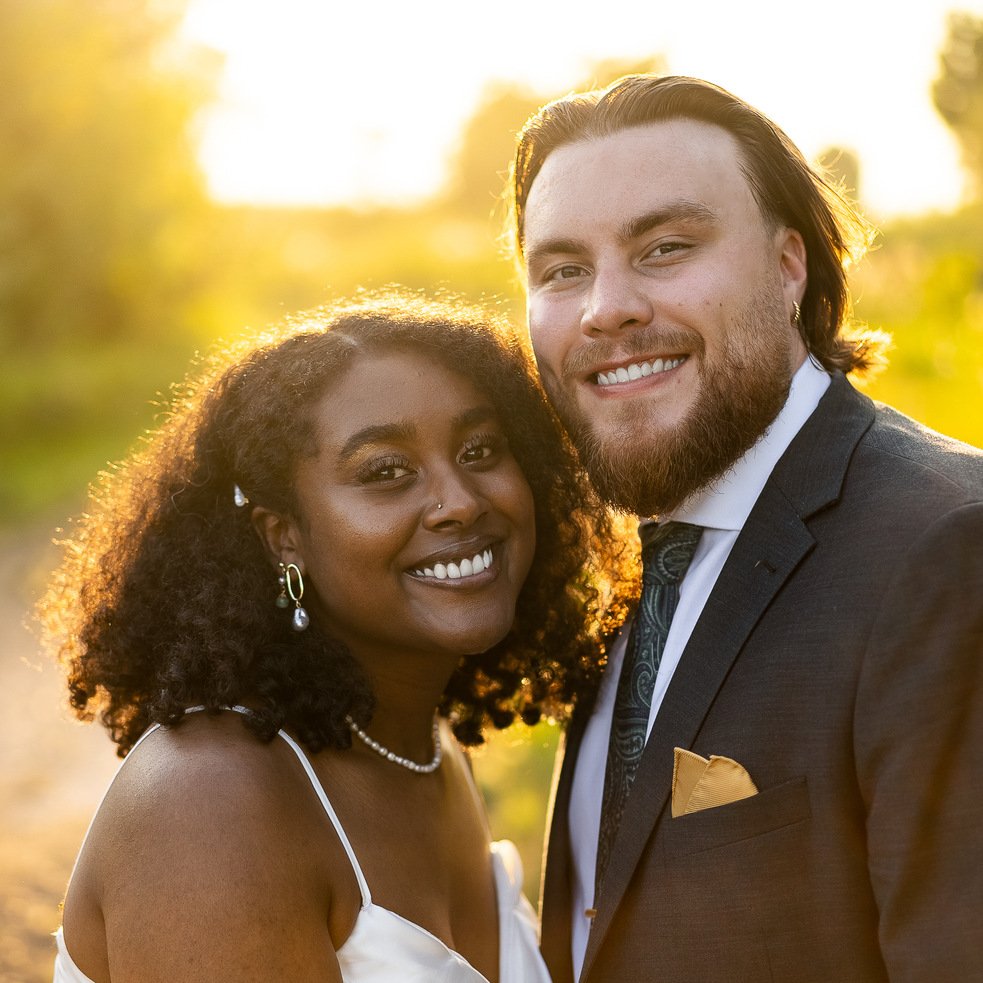 A smiling couple dressed in formal attire outdoors during golden hour, with the woman wearing a white dress and jewelry, and the man in a dark suit with a pocket square, near sunset with trees in the background.