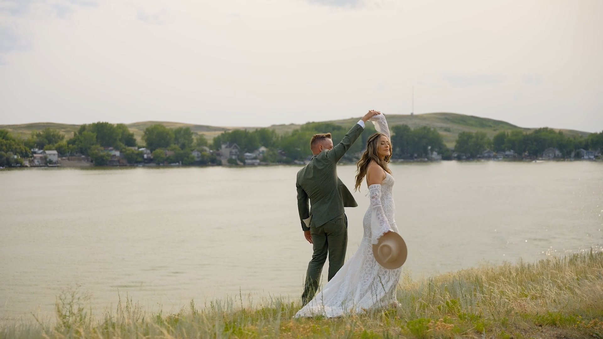 A couple in wedding attire dancing by a lake, with the man twirling the woman, holding her hand and her dress, as she holds a hat in her other hand, outdoors near grassy banks.