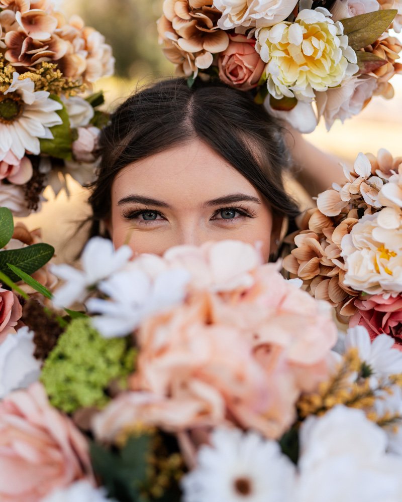 A woman with blue eyes and dark hair surrounded by a variety of colorful flowers, peeking over them.