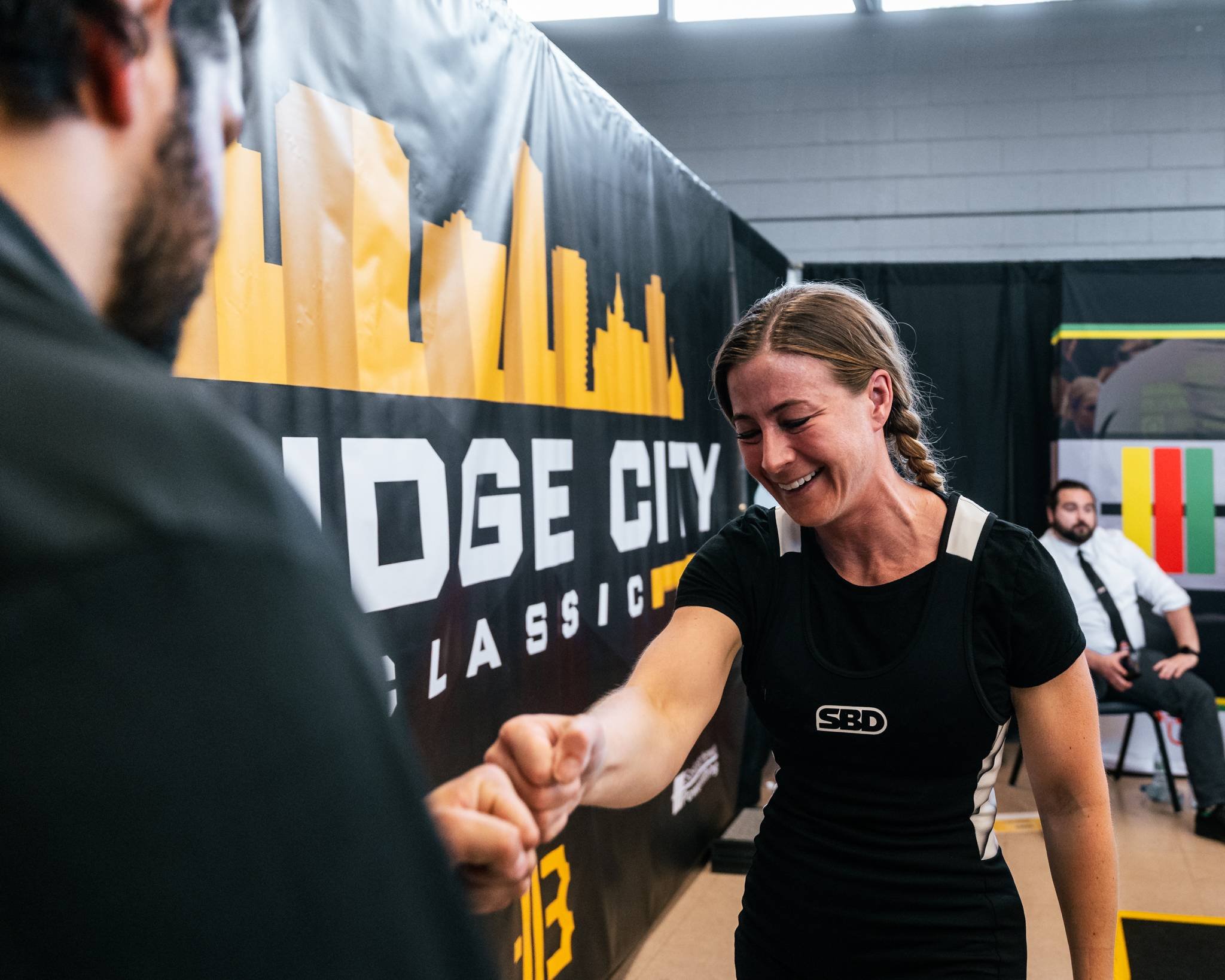 A woman in athletic clothing shaking hands with a man at a fitness event, with a backdrop reading 'Edge City' and a seated man in the background.