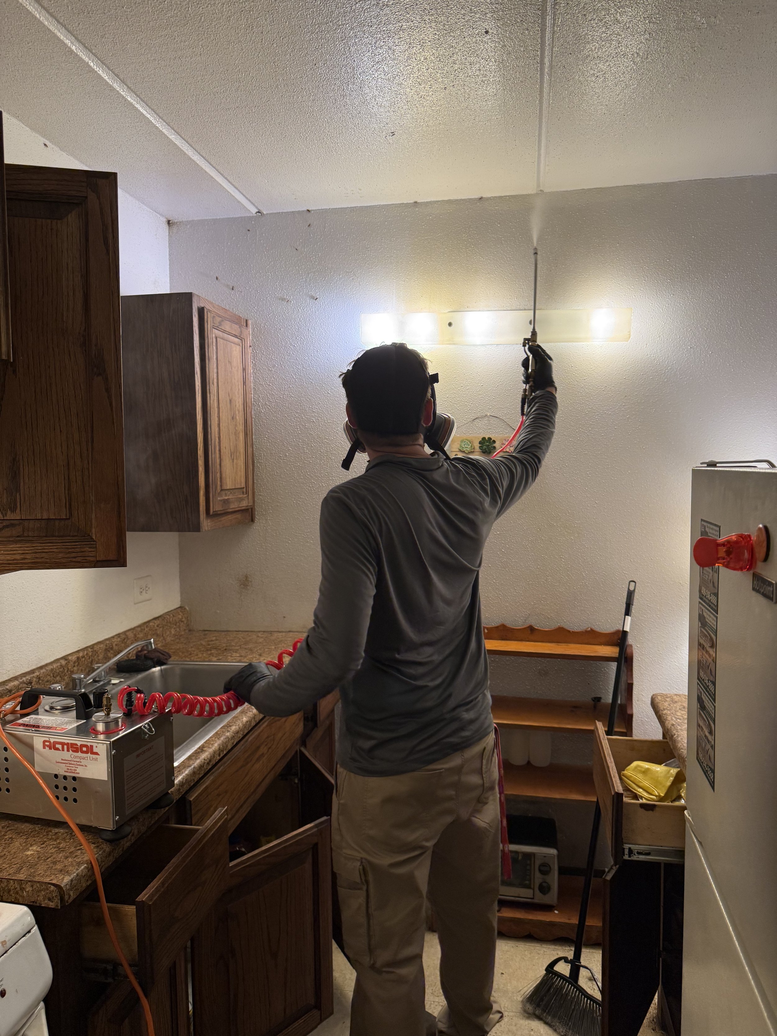 A person wearing safety goggles and a mask is spraying a kitchen ceiling with a cleaning or disinfecting solution using a spray gun, standing in front of kitchen cabinets and counter.
