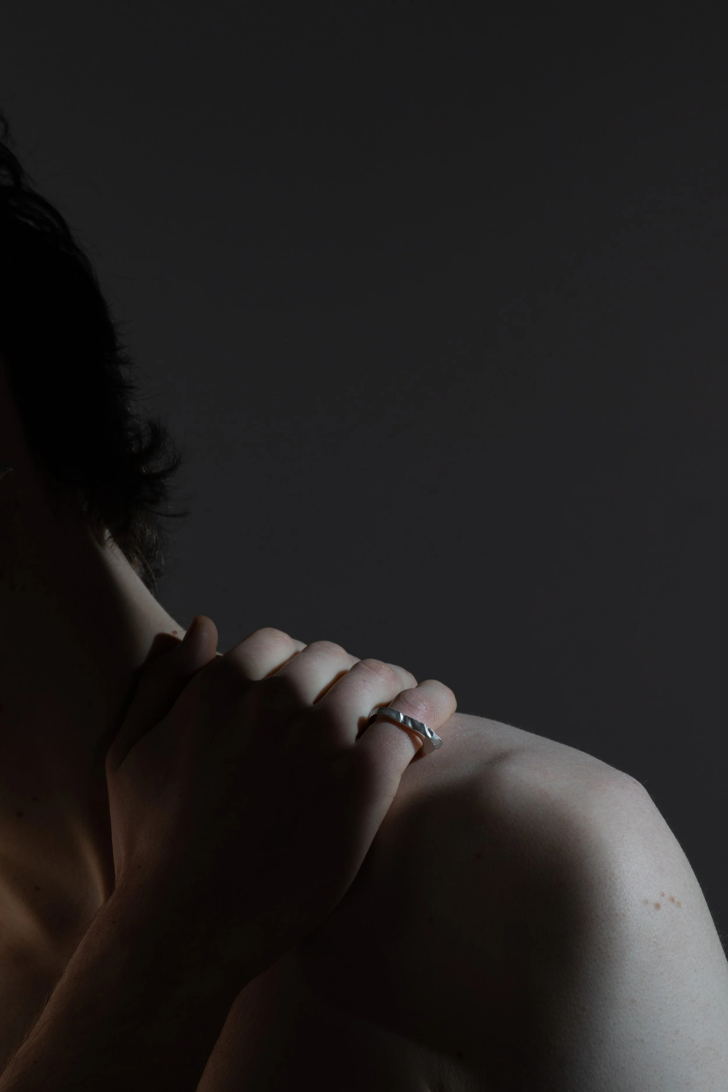 Close-up of a person's hand resting on their shoulder with a silver ring visible, soft lighting creating shadows on the skin.