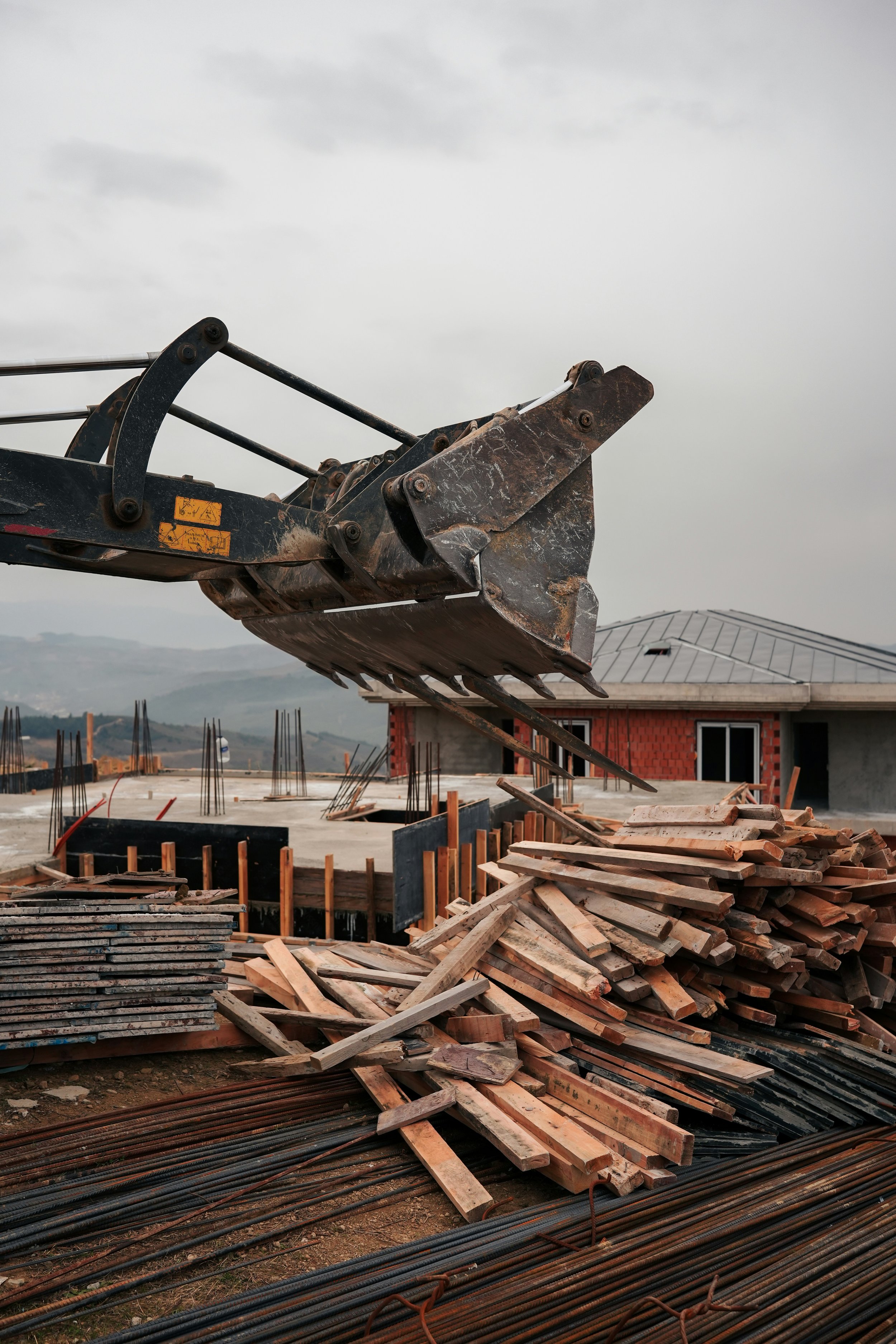 Baustelle mit Baugerät, Holzbohlen und Stahlträger, im Hintergrund ein Haus mit Metall-Dach, bewölkter Himmel.