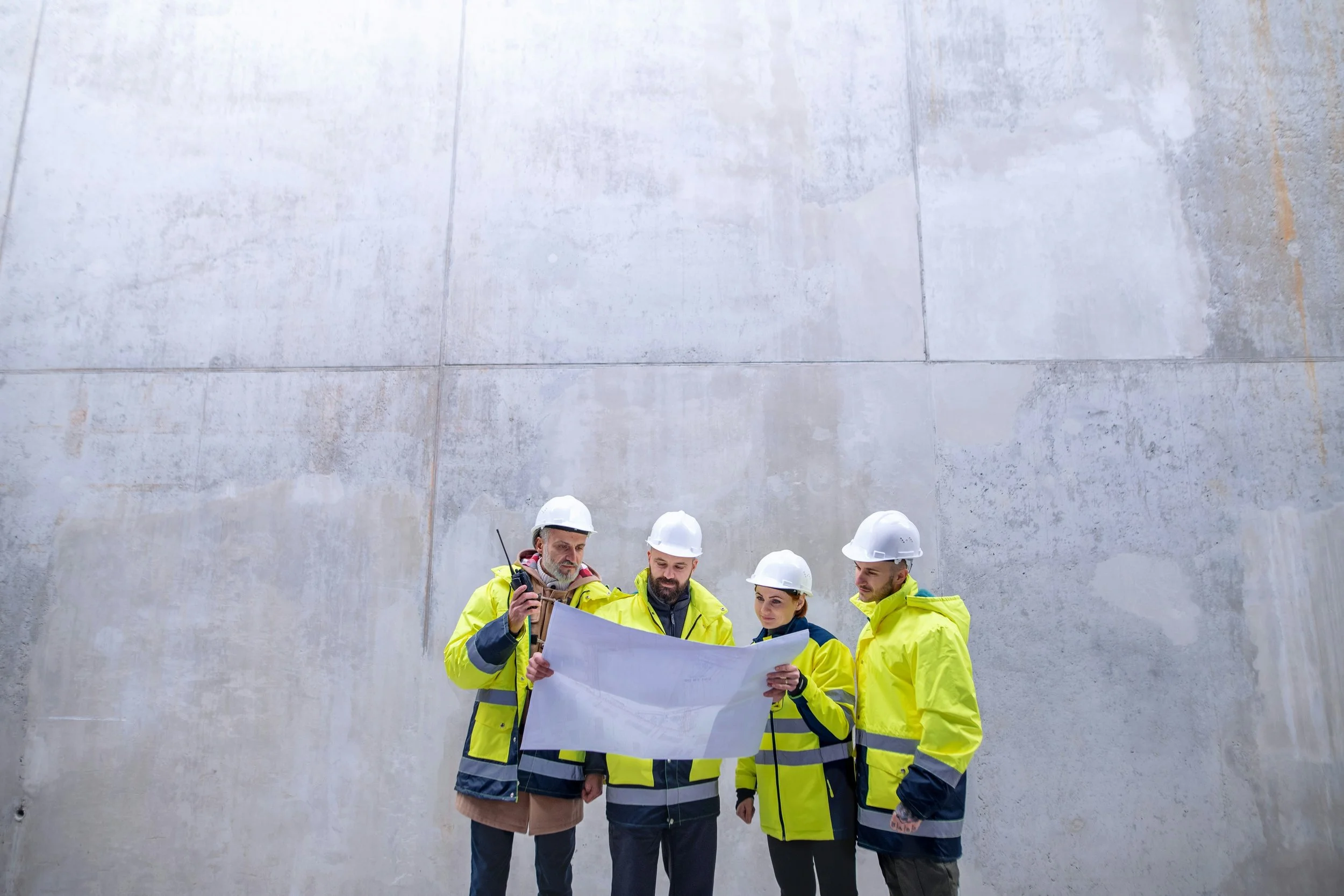 Vier Bauarbeiter in gelben Sicherheitswesten und weißen Helmen stehen vor einer Betonwand und schauen sich einen Bauplan an.