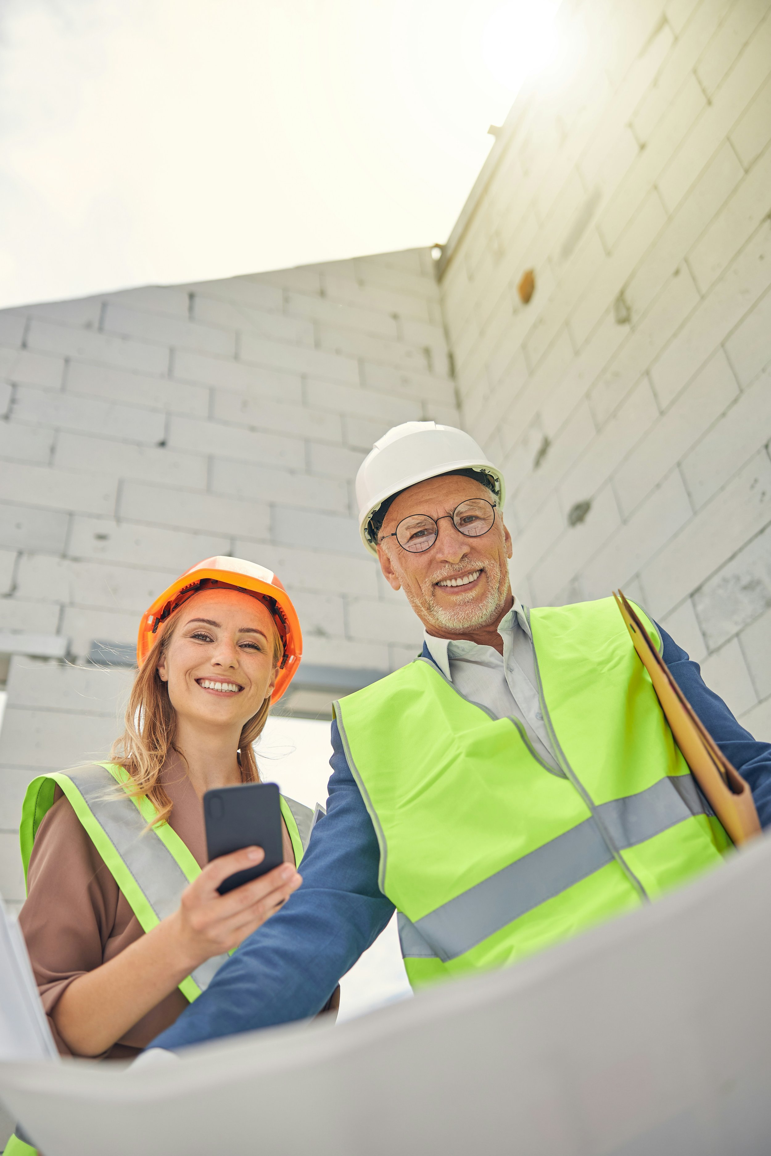Zwei Bauarbeiter mit Schutzhelmen und gelben Sicherheitswesten auf einer Baustelle, lachend und ein Smartphone betrachtend.