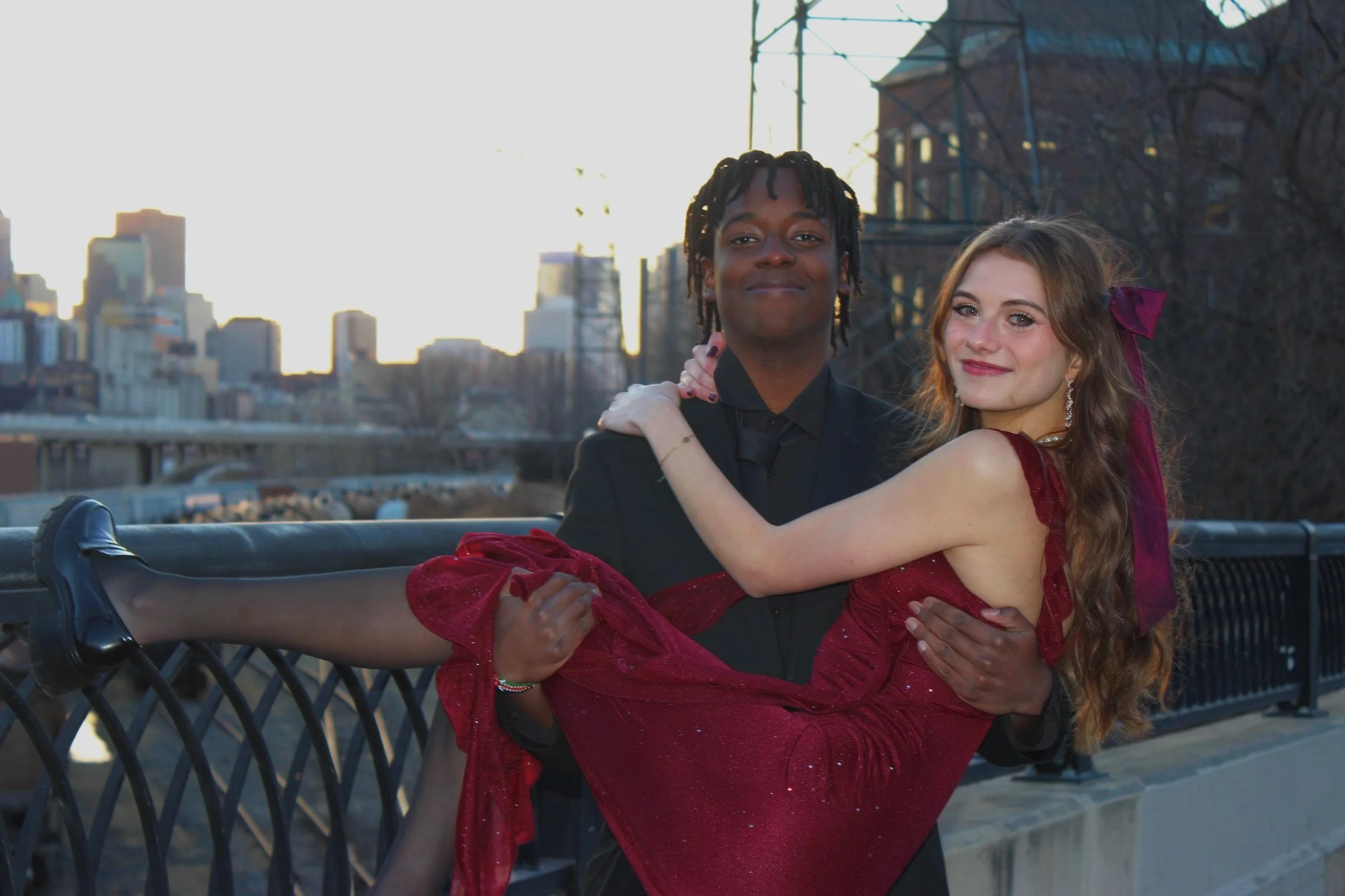 A young couple poses on a city bridge during sunset, with the man carrying the woman in a red dress. The city skyline is visible in the background.