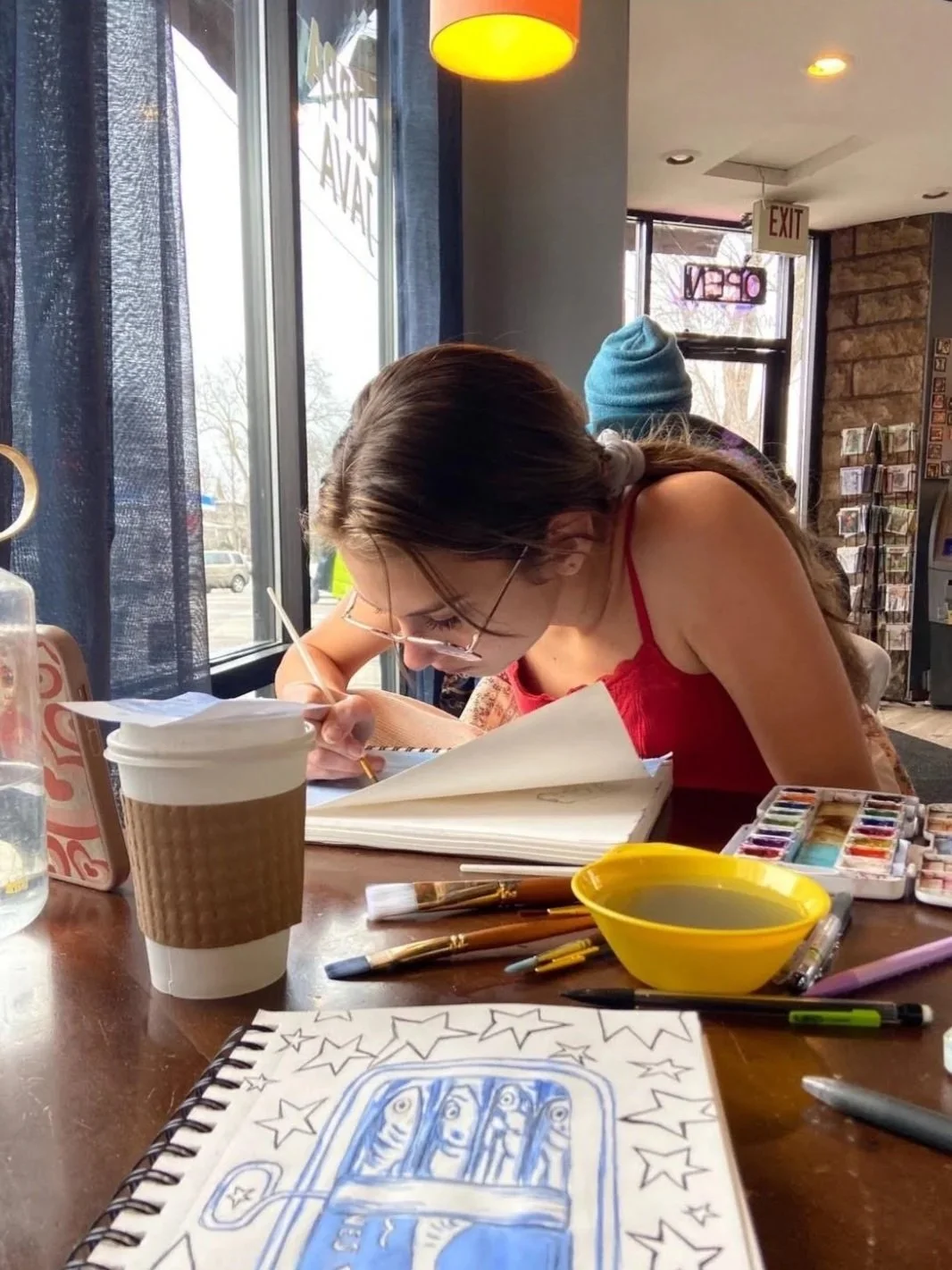 A woman with long brown hair, wearing glasses and a red tank top, is sitting at a table in a cafe, drawing or writing in a sketchbook. Various art supplies, including paintbrushes, watercolors, pens, and a yellow bowl, are spread out on the table. There is a disposable coffee cup in front of her, and a bottle of water on her left. The cafe has large windows and a pack of paper with star and face drawings is visible in the foreground.