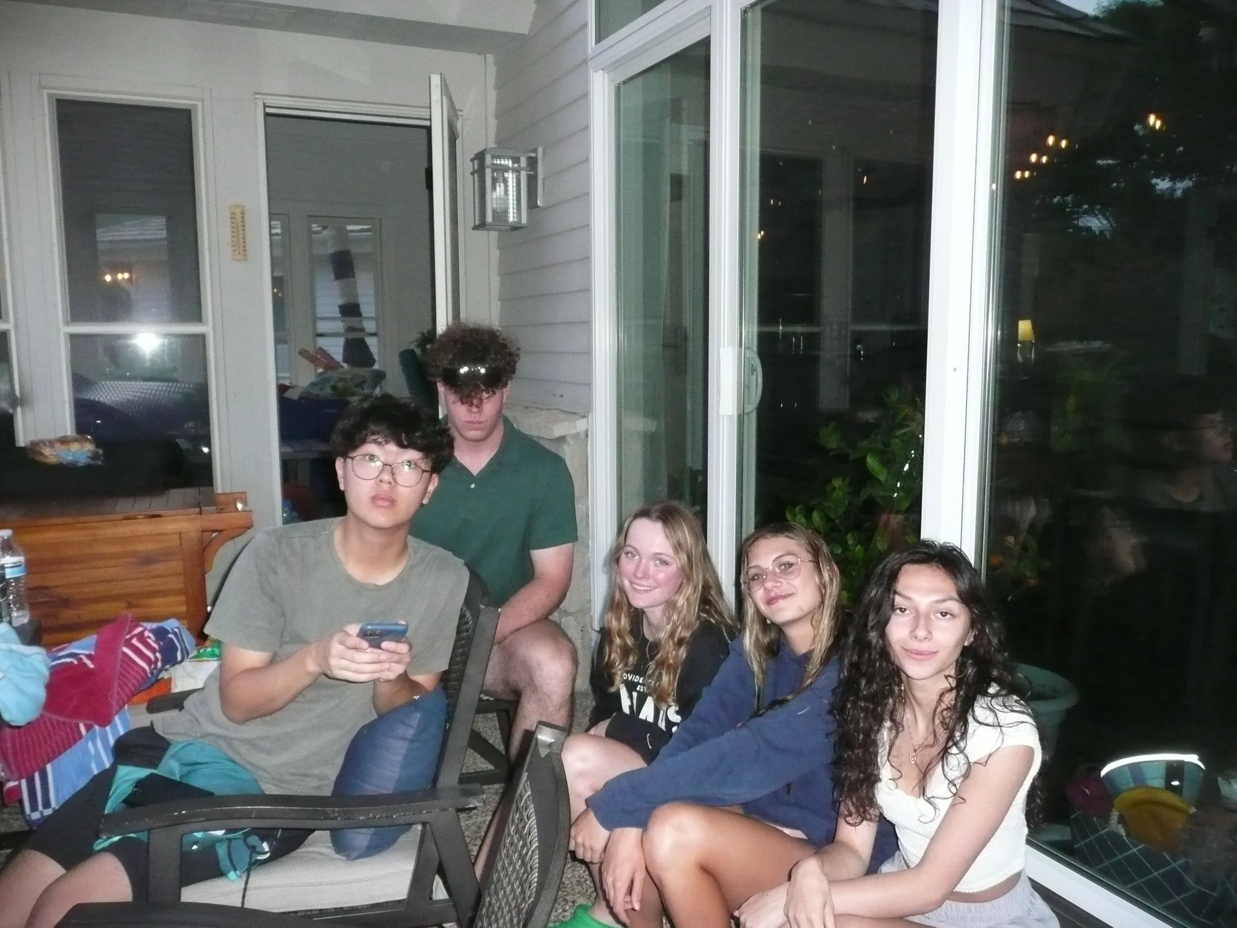 Group of five young friends sitting close together on a porch or patio at night, some smiling and looking at the camera, one person looking at a phone, with a glass door and windows behind them.