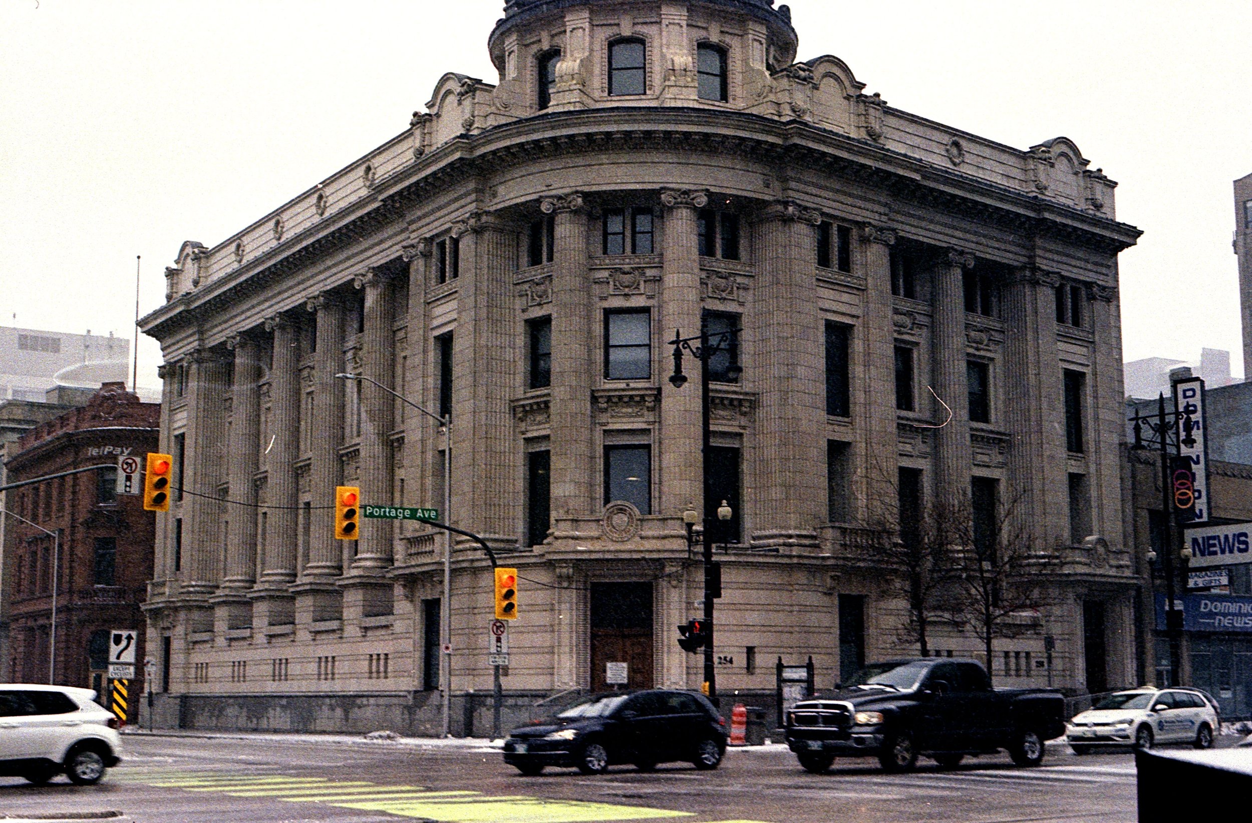 A historic stone building with multiple columns and ornate architectural details at the corner of a city street, with a street sign reading 'Portage Ave' and traffic lights, and cars driving by in the foreground.