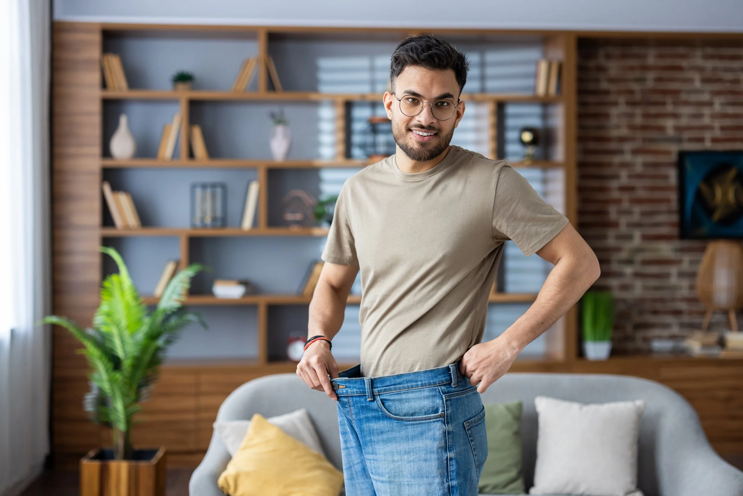 Young man with glasses smiling and holding out the waist of his jeans to show they are too big, in a modern living room.
