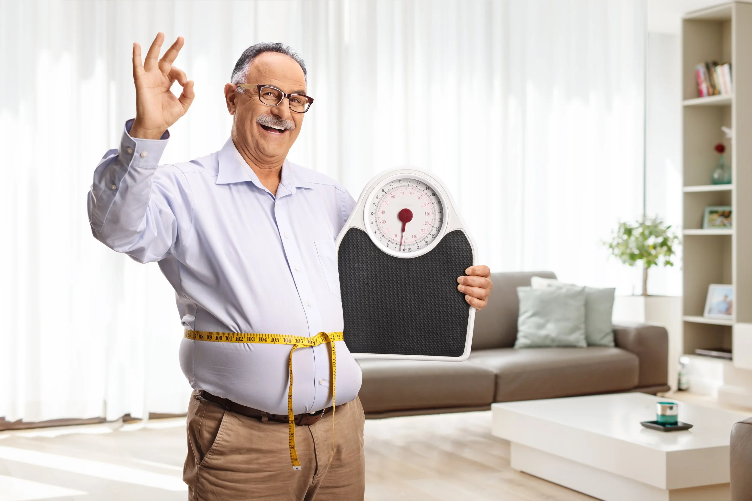 An elderly man smiling and making an 'OK' gesture with his right hand, holding a digital bathroom scale, standing in a bright living room with a sofa and bookshelf in the background, with a measuring tape around his waist.