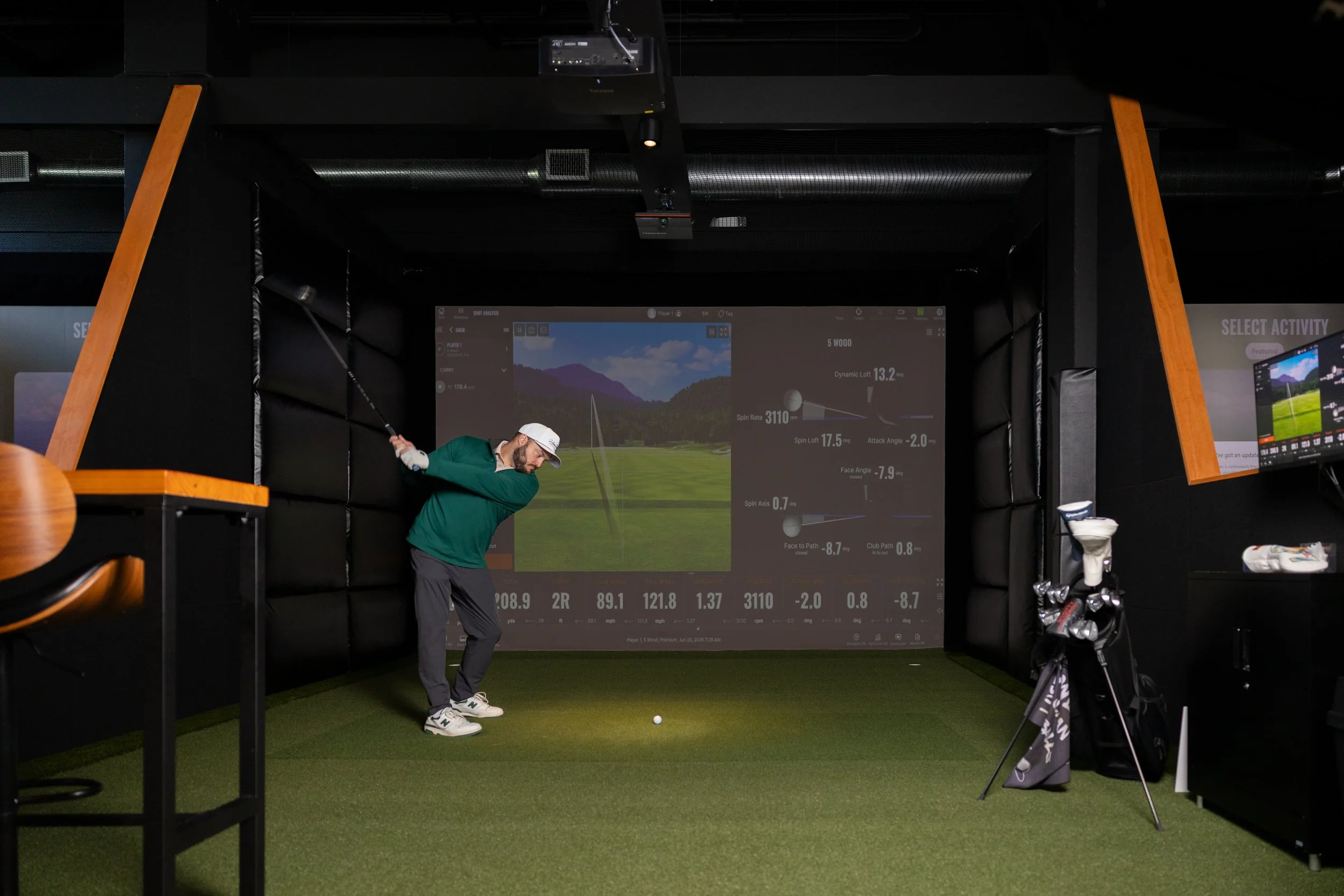 Man practicing golf swing indoors in a golf simulation room with a large screen displaying a golf course and shot data.