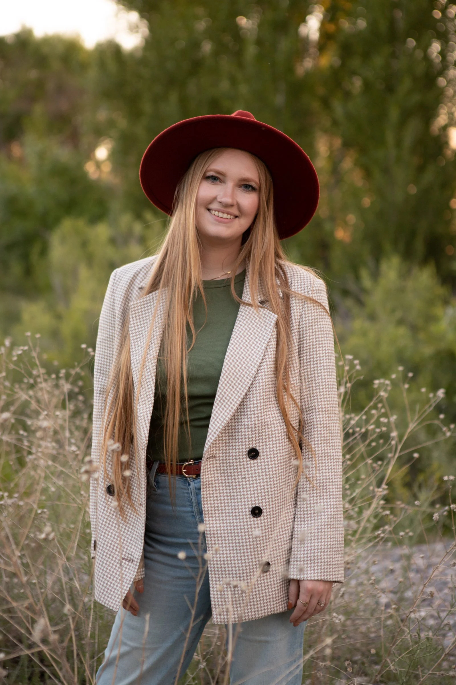 A young woman with long red hair, wearing a wide-brimmed red hat, a checked blazer, a green top, and blue jeans, standing outdoors in a field with trees in the background.