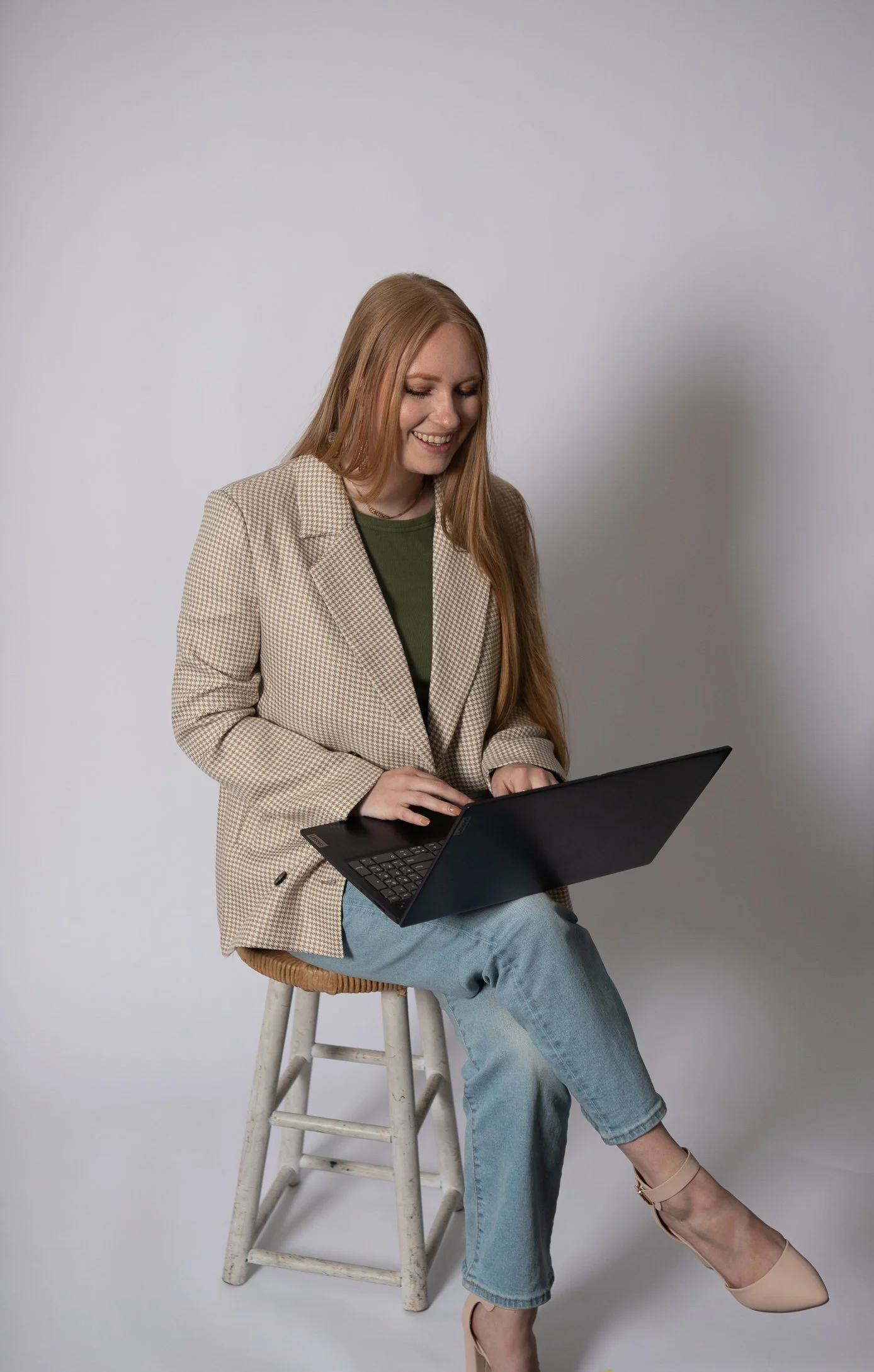 A woman with long red hair sits on a stool, smiling and using a laptop against a plain light gray background.