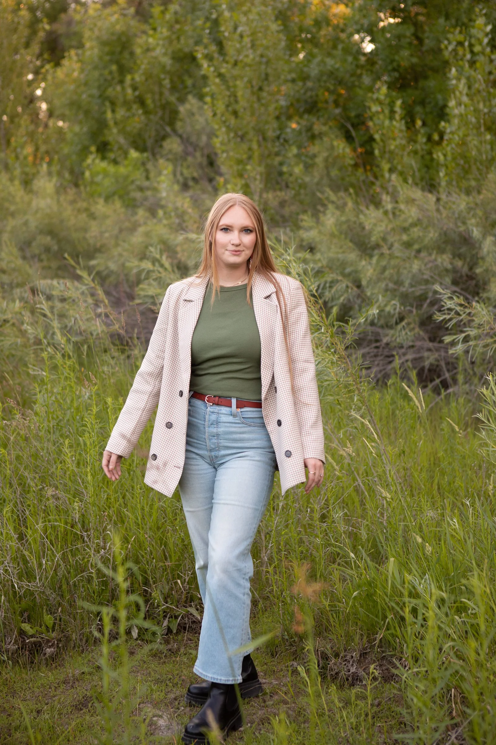 A young woman walking through a grassy area with trees in the background, wearing a beige checkered coat, green shirt, light blue jeans, and black boots.