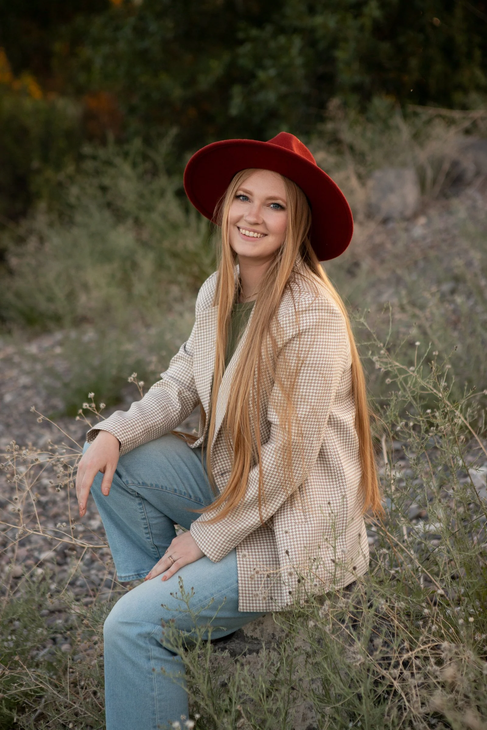 A woman with long red hair, wearing a beige checked blazer, blue jeans, and a large red wide-brimmed hat, smiling while sitting outdoors on rocky terrain surrounded by sparse plants and greenery.