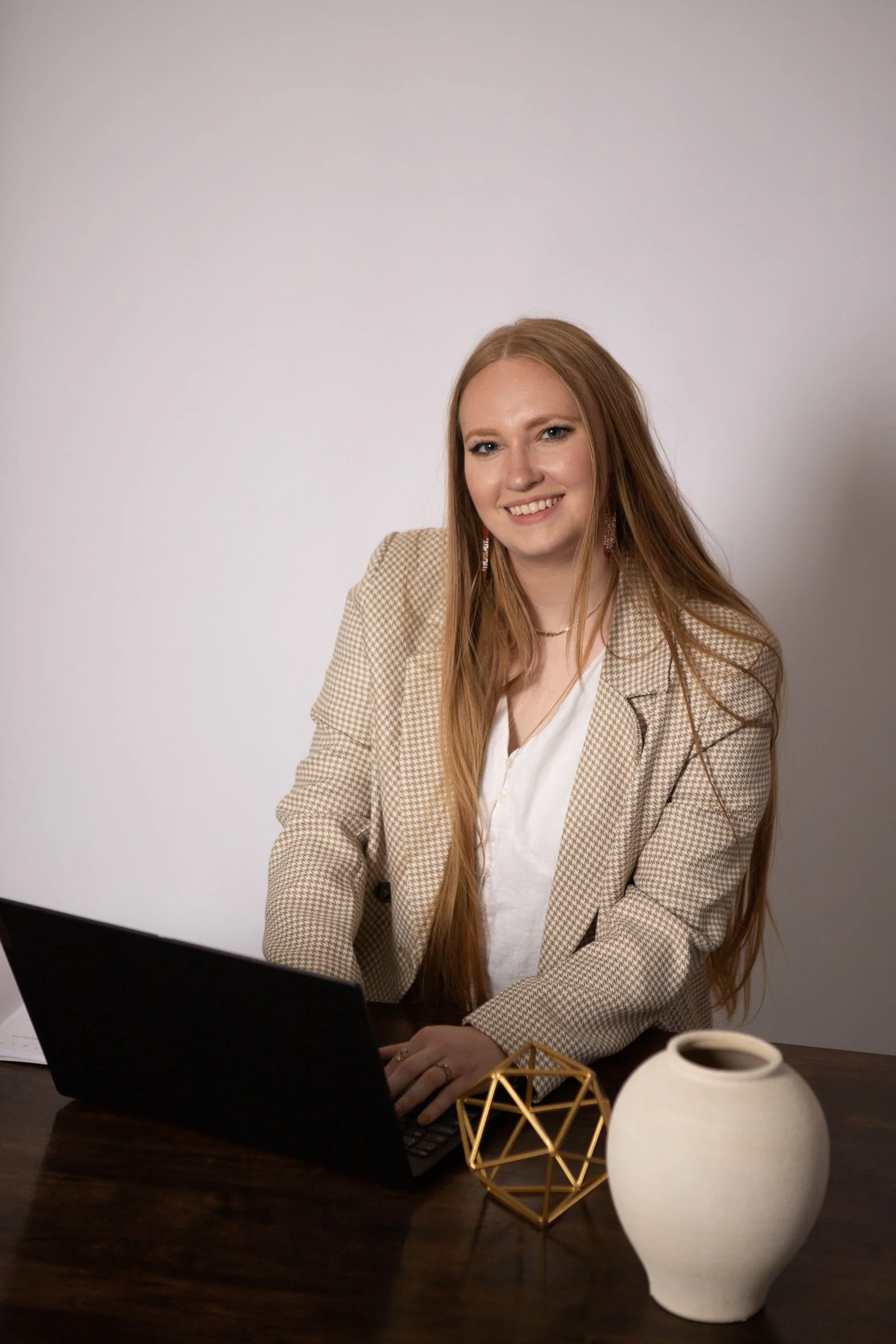 A young woman with long red hair sitting at a desk with a laptop, a geometric gold decor piece, and a white ceramic vase, smiling at the camera.