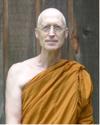 Bald man wearing glasses and an orange Buddhist monk robe standing in front of a wooden fence.