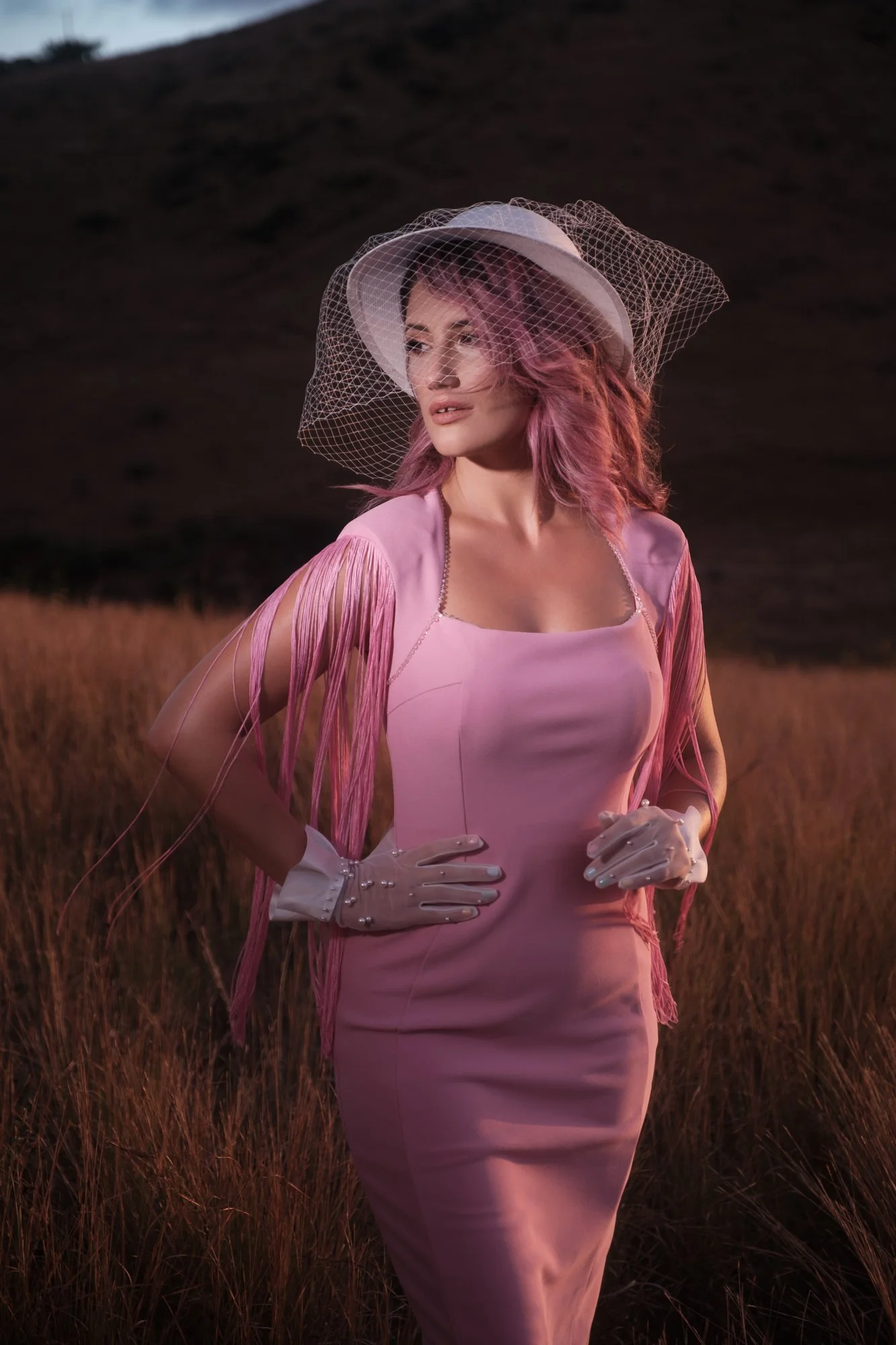 Femme en robe rose, chapeau blanc avec voile, dans un champ au coucher du soleil.