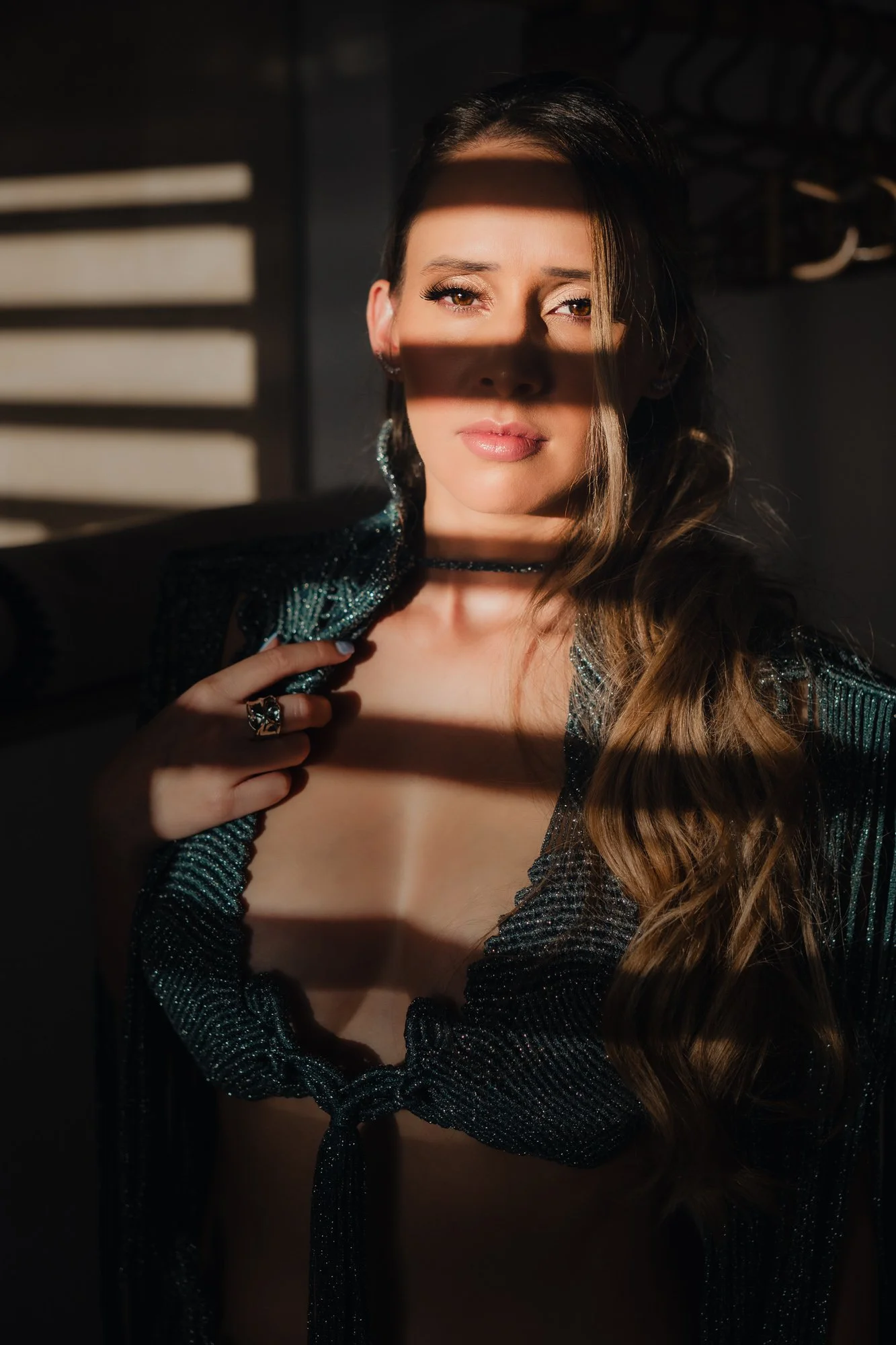 Une femme aux longs cheveux châtains, portant une robe en macramé brillante verte décolletée, se tient devant une fenêtre éclairée par le soleil créant des ombres sur son visage.