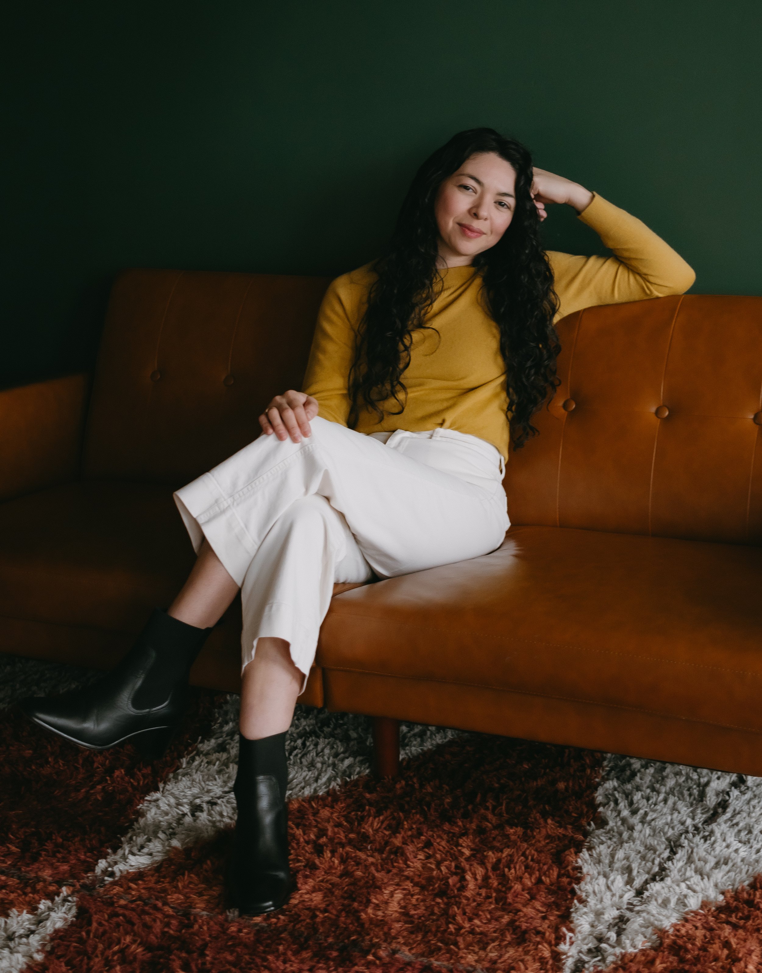 A woman with long curly hair sitting on a brown leather couch, wearing a mustard yellow top, white pants, and black ankle boots, winking at the camera, against a dark green wall.