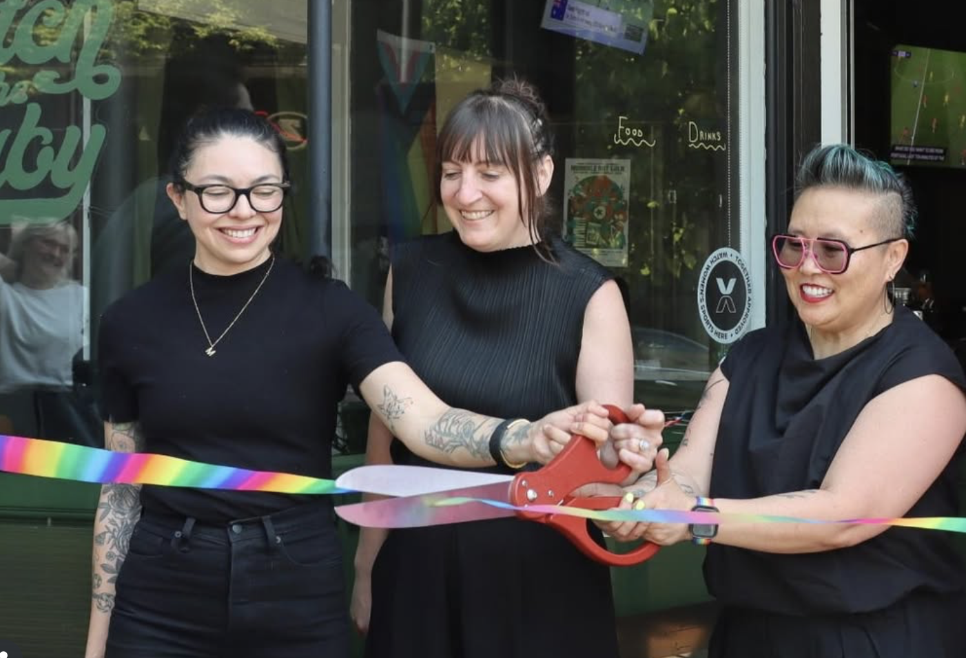 Three women participating in a ribbon-cutting ceremony, with one woman holding large scissors and cutting a rainbow-colored ribbon in front of a storefront window.