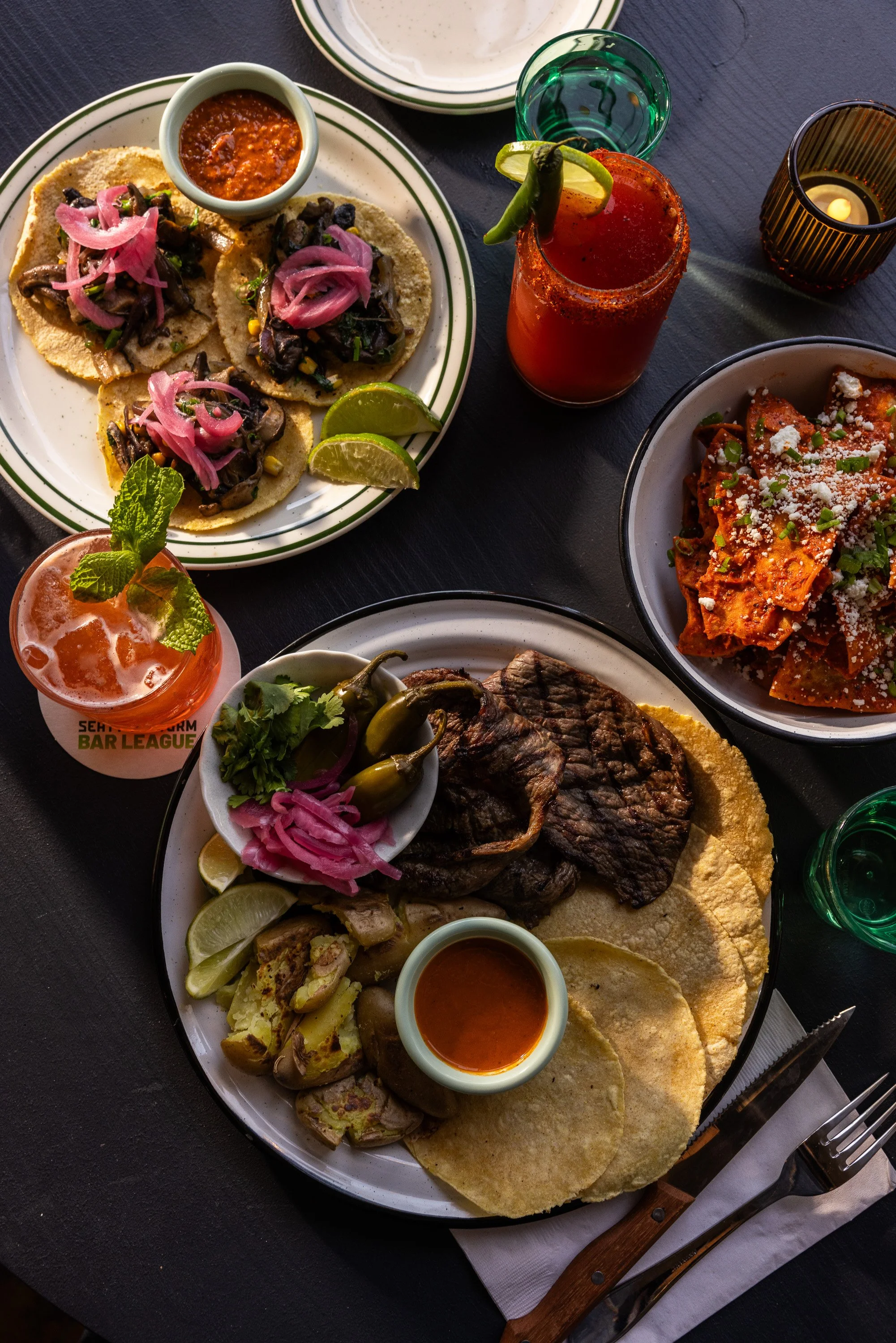 A Mexican meal featuring tacos with pickled pink onions, lime wedges, grilled steak, rice, and a side of pasta with cheese, served with cocktails and lime drinks on a dark table.