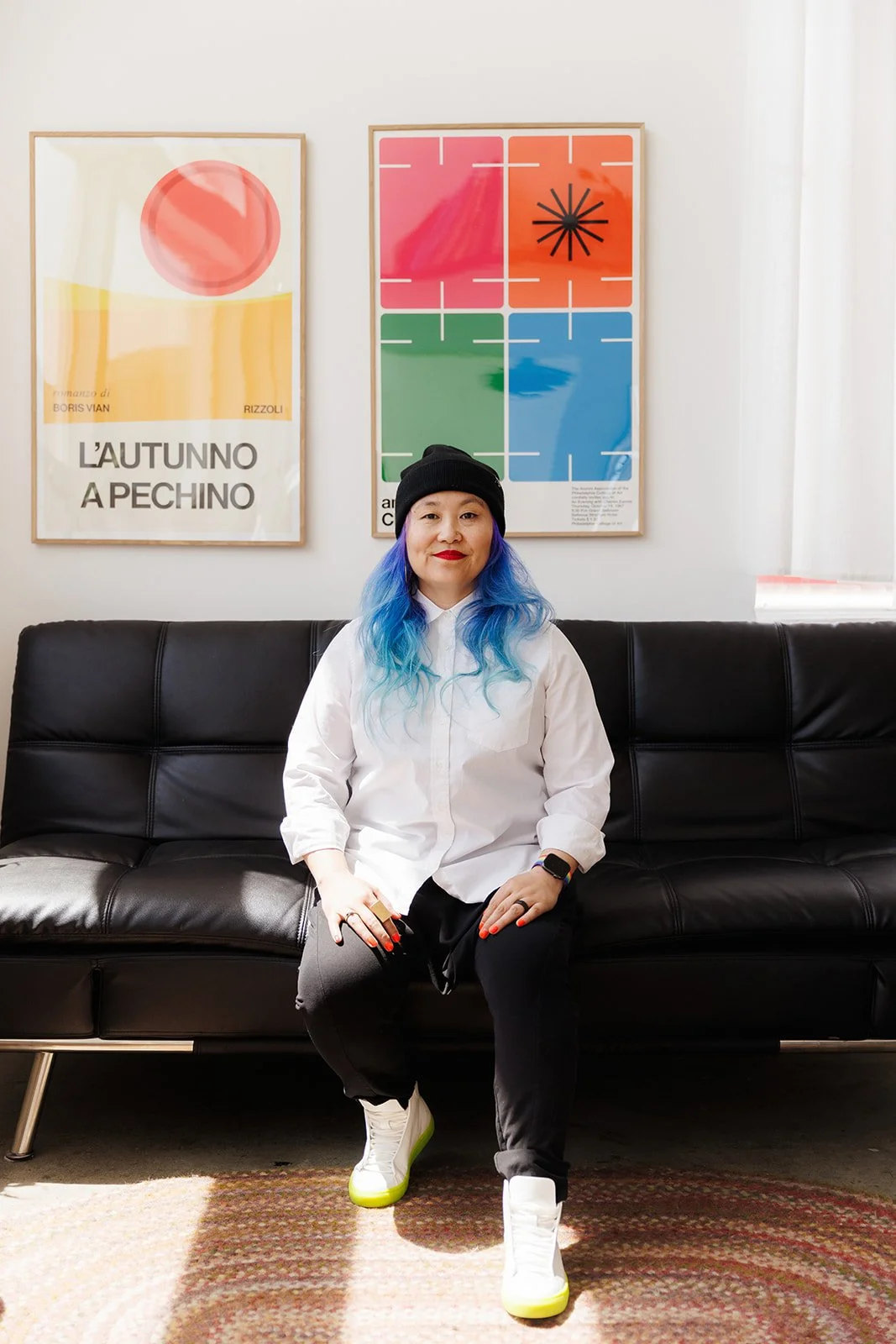 A woman with blue hair seated on a black leather couch in a room with white walls. She is wearing a black beanie, a white shirt, black pants, and white sneakers with green soles. Behind her, there are two colorful framed art posters on the wall.