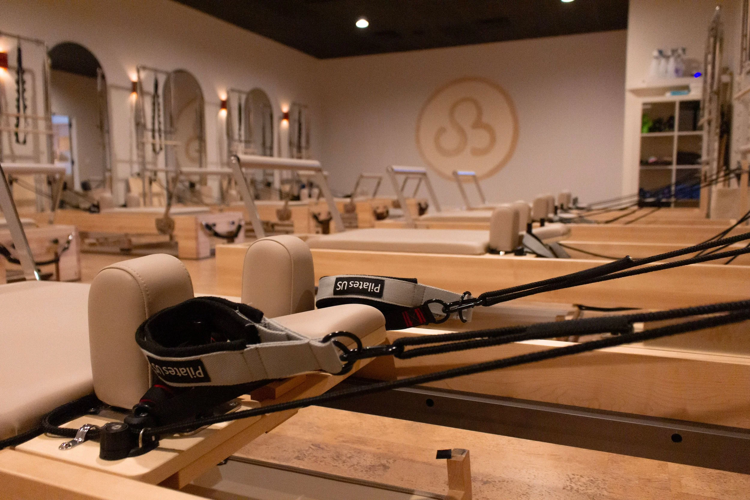 Pilates reformer machines in a yoga or Pilates studio with beige upholstered seats, ropes and pulleys, and a logo on the wall in the background.