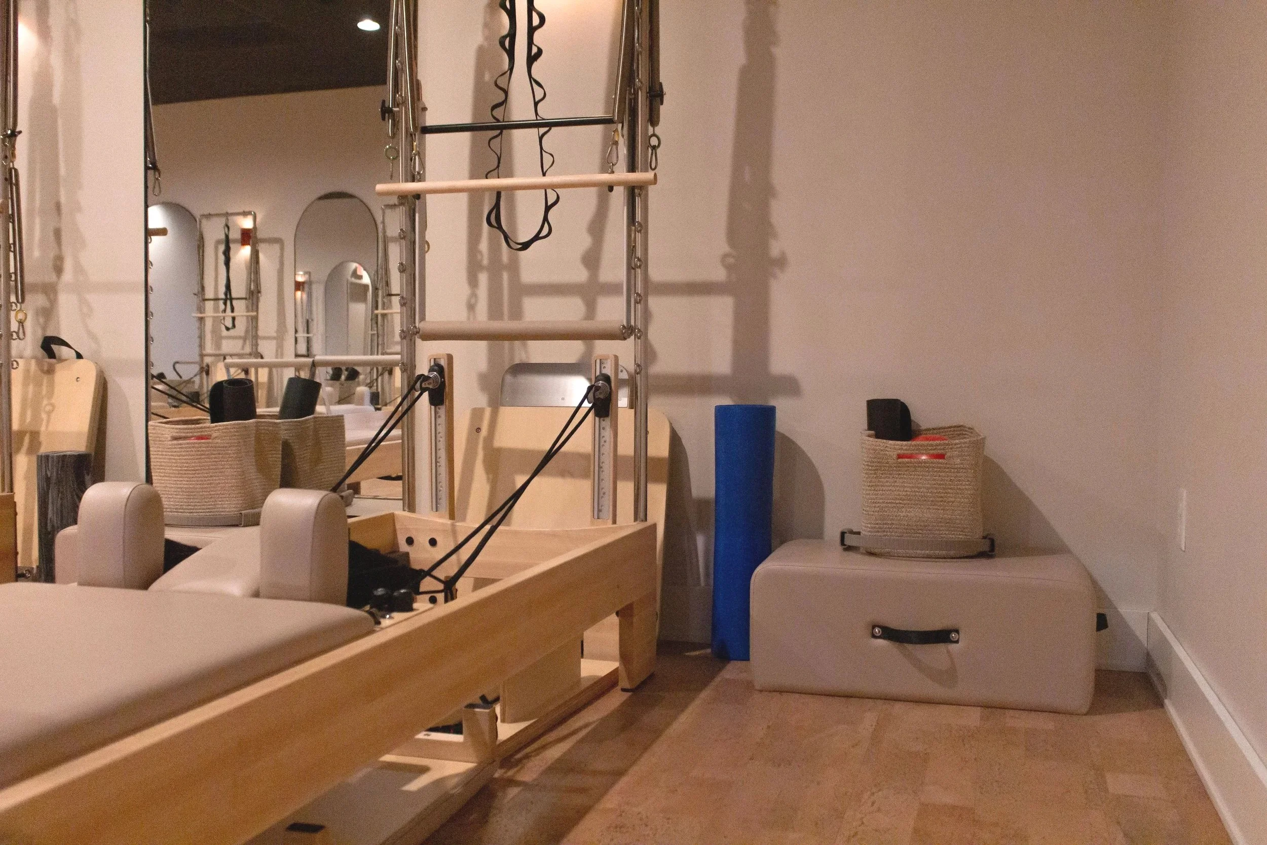 A Pilates reformer machine and a padded mat with a blue foam roller, as well as storage baskets, are visible in a room with mirrors and beige walls.