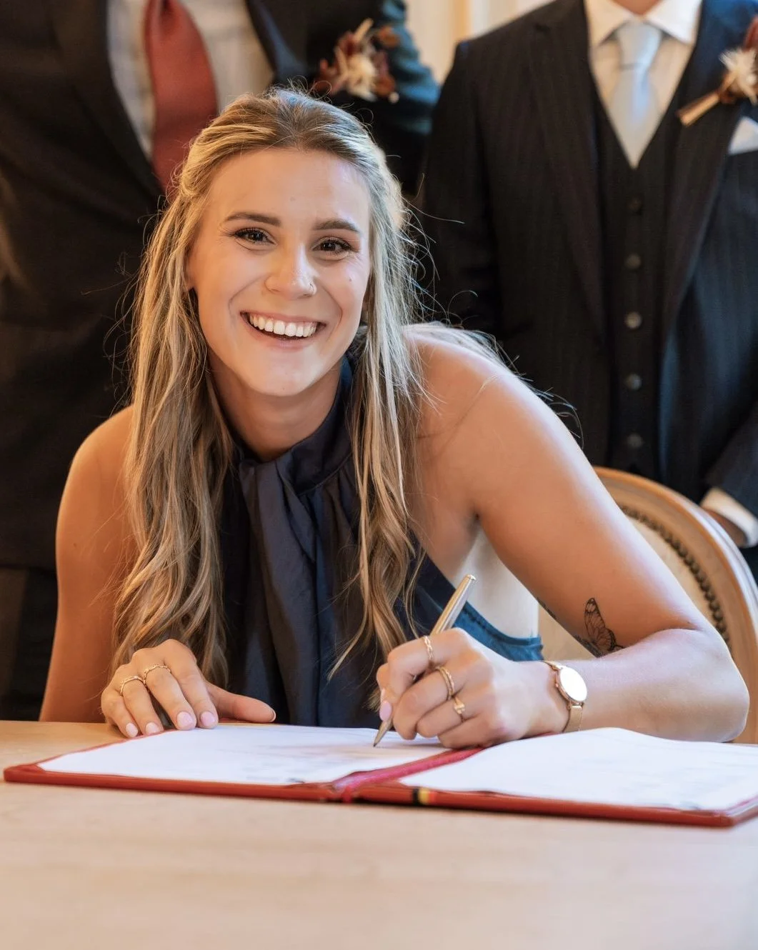 A young woman with long blonde hair smiling while signing a document at a formal event, wearing a sleeveless black dress and gold jewelry.