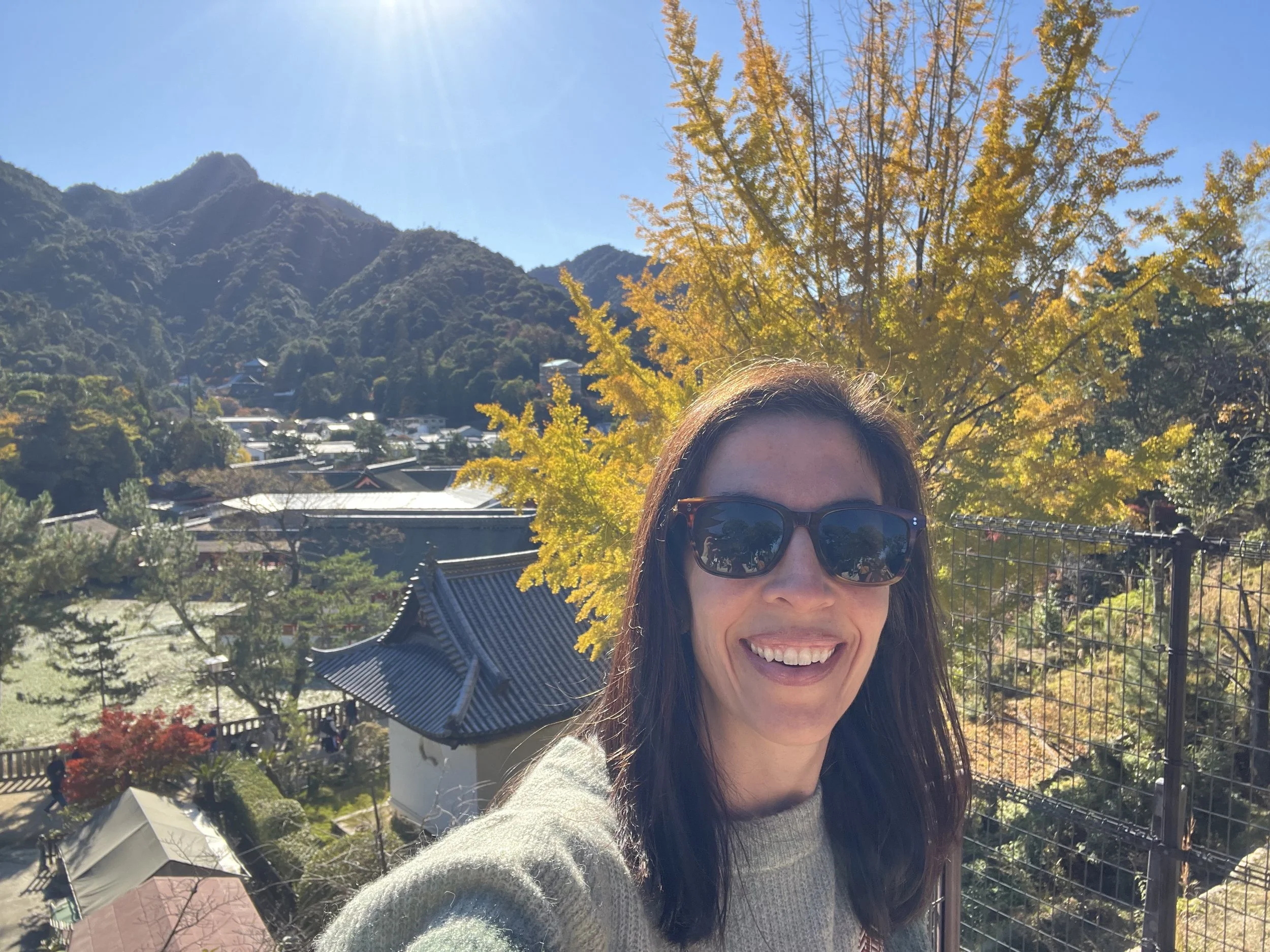 Smiling woman taking a selfie outdoors with mountains, a yellow autumn tree, traditional buildings, and a clear blue sky in the background.