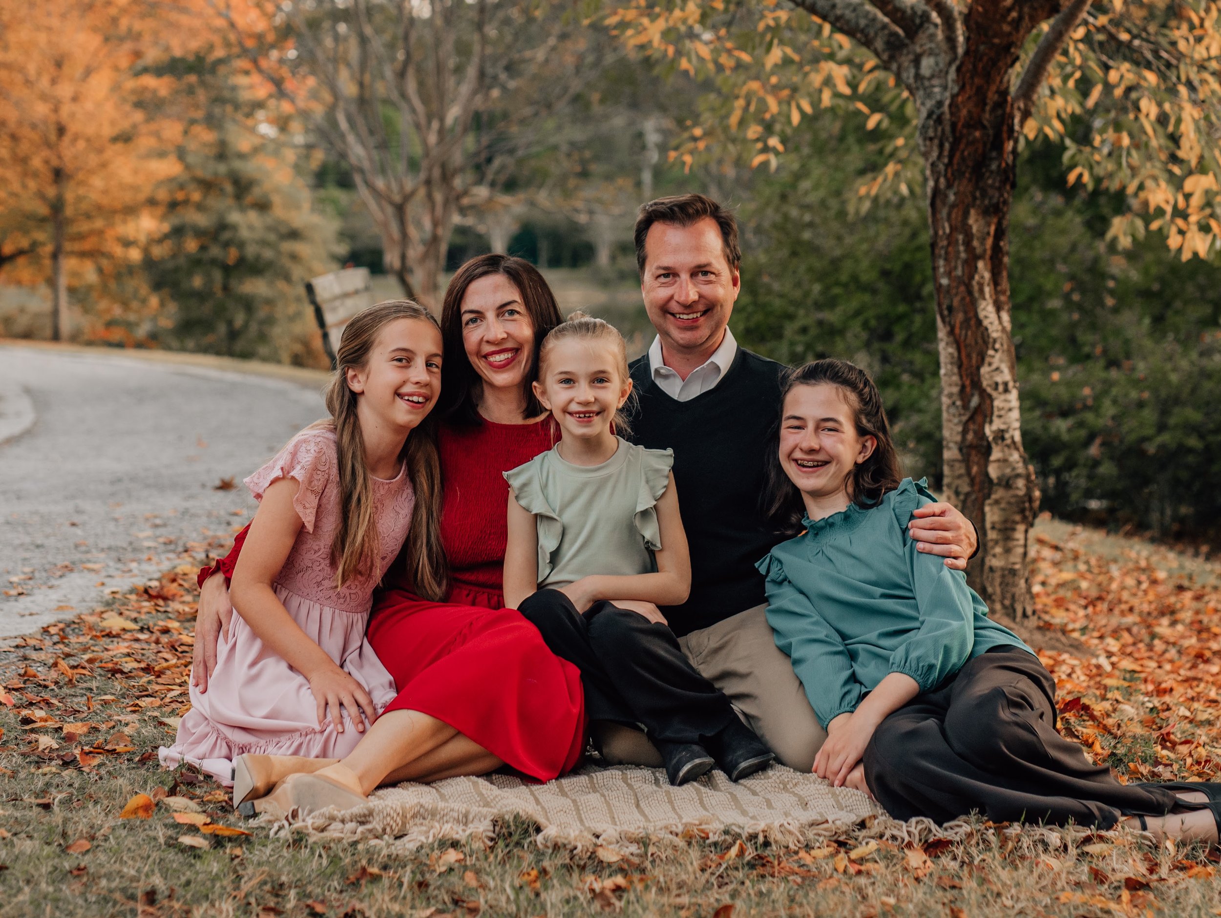 A family of six, including two parents and four children, sitting on a blanket outdoors during autumn, surrounded by fall-colored trees.