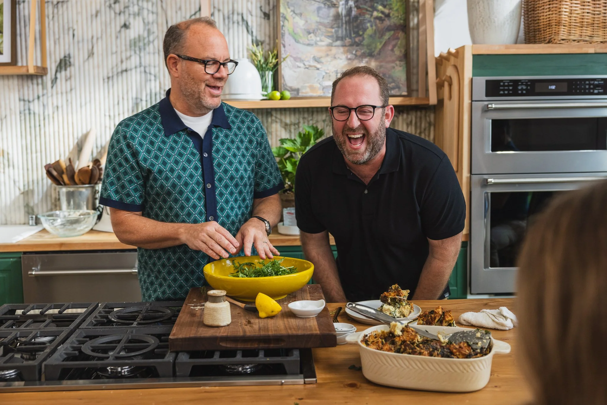 Two men are laughing and enjoying a moment in a kitchen, with one preparing a salad and the other with leftover food on the counter.