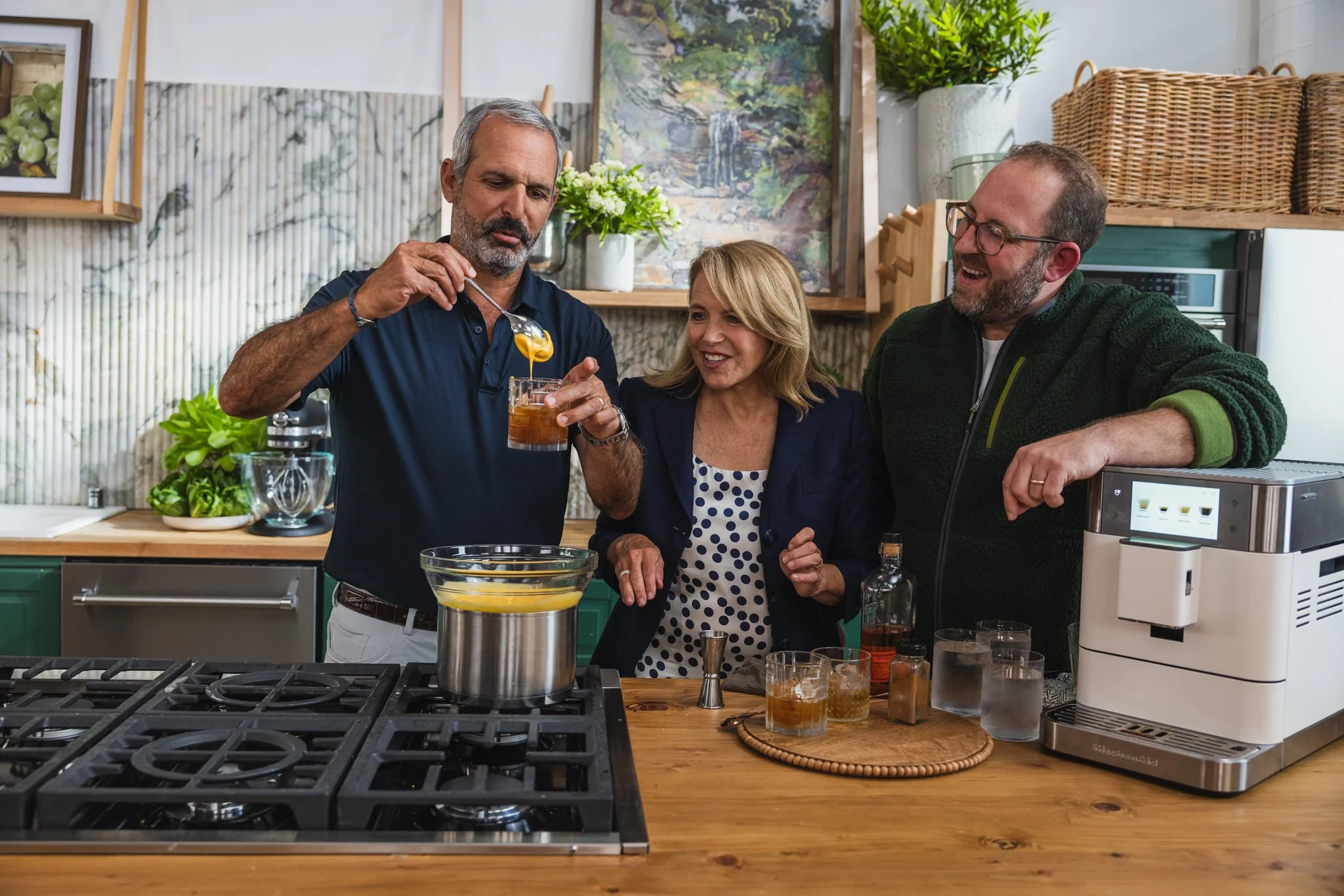 Three adults in a kitchen, pouring drinks and chatting, with various kitchen items and plants in the background.