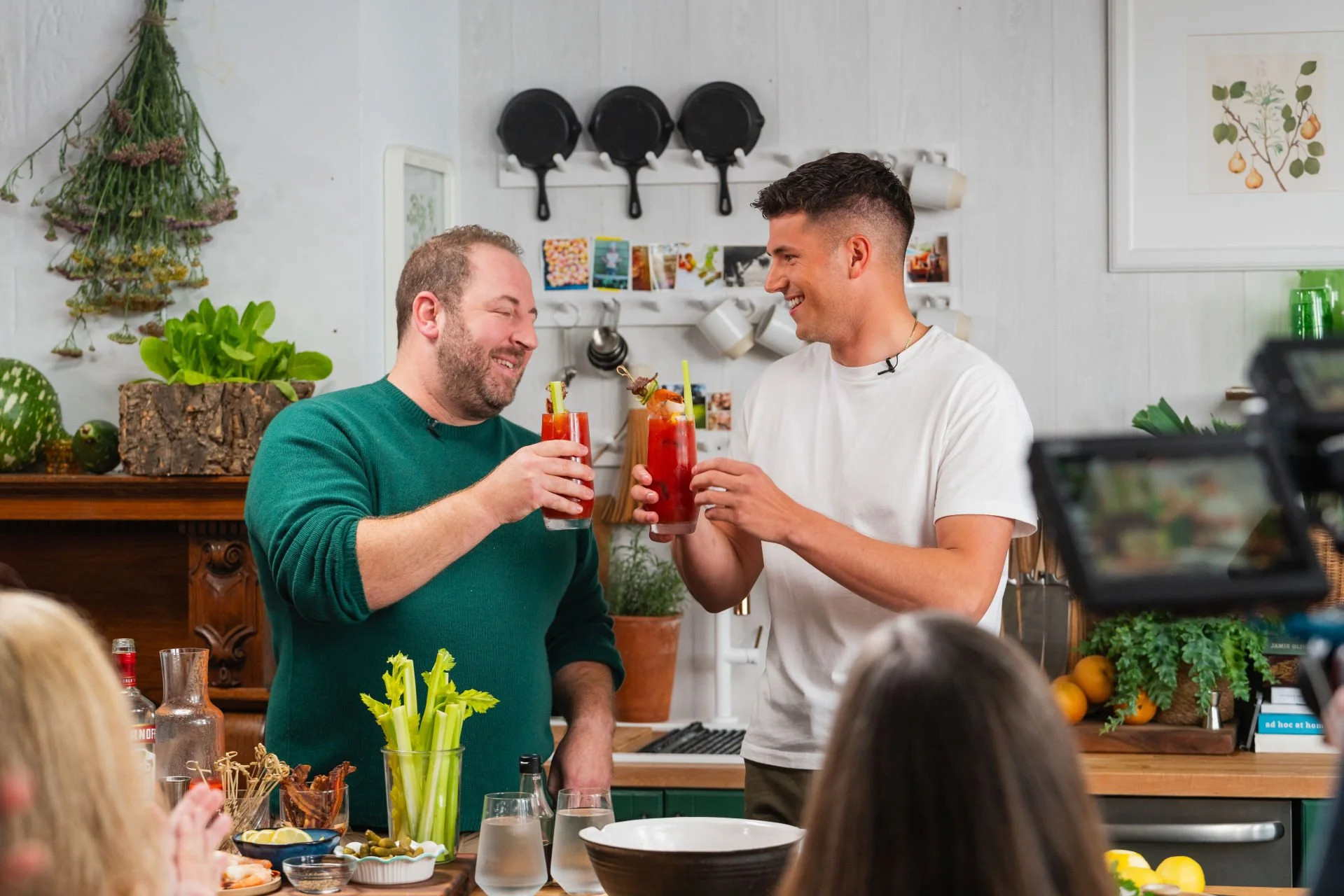 Two men sharing a toast with Bloody Mary cocktails in a cozy kitchen during a gathering.