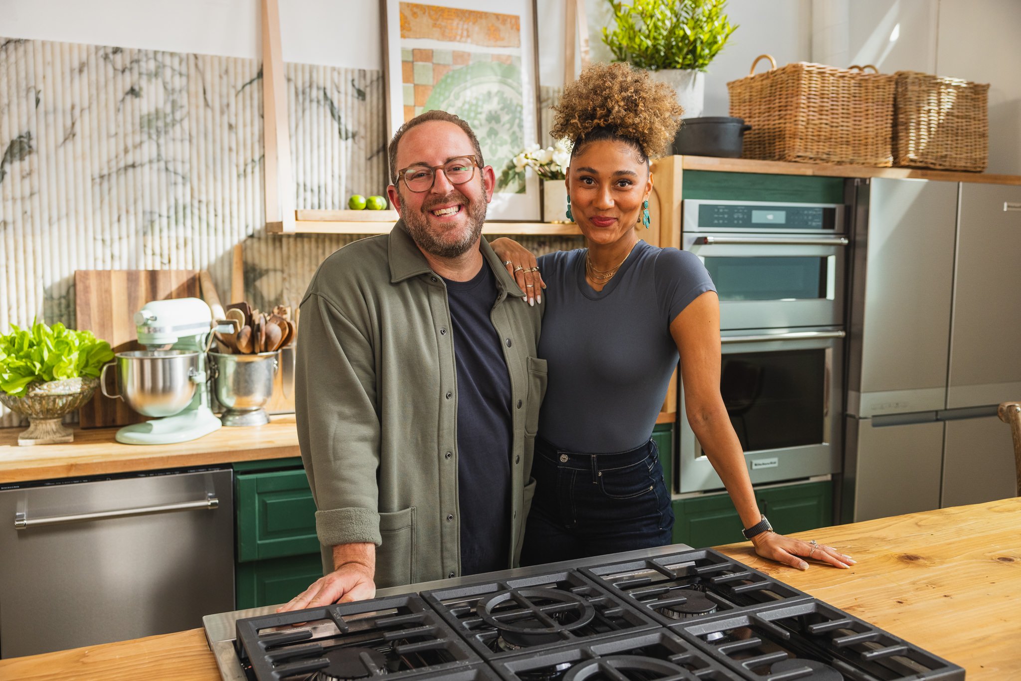 A smiling man and woman standing in a modern kitchen, leaning on a wooden countertop near a gas stove, surrounded by kitchen appliances, plants, and artwork.