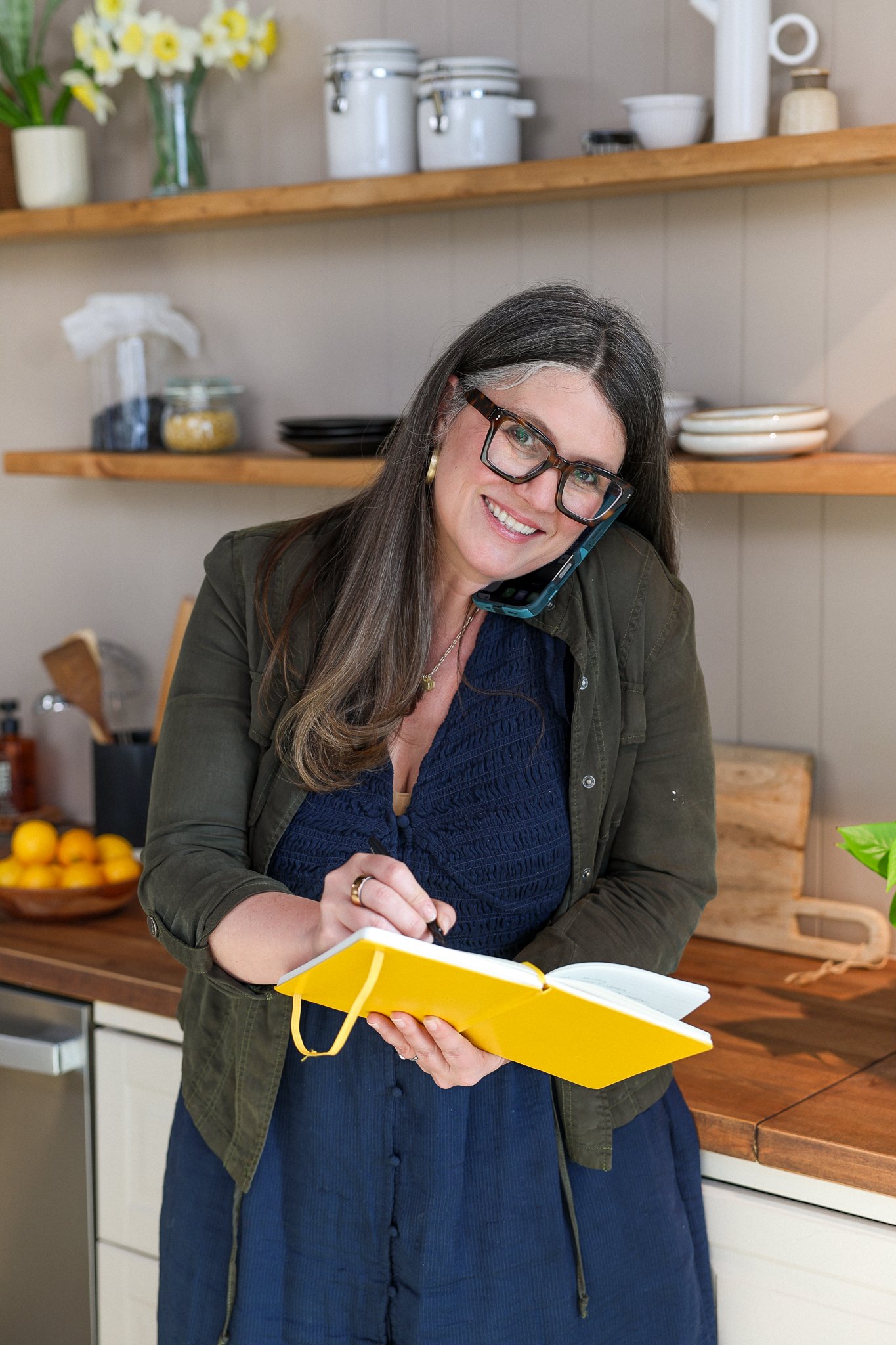 A woman with long wavy hair, glasses, and a black blazer smiling in a kitchen with colorful artwork and shelving in the background.