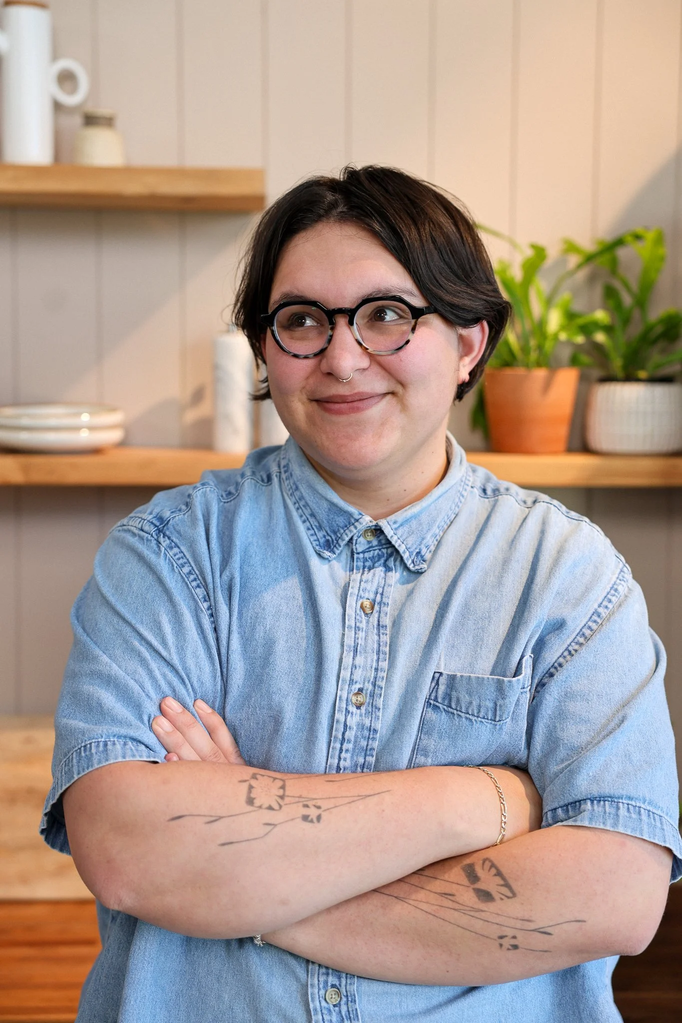 A smiling person with short dark hair, round glasses, and a septum piercing, wearing a black button-up shirt, standing outdoors under a structure with blurred city background.