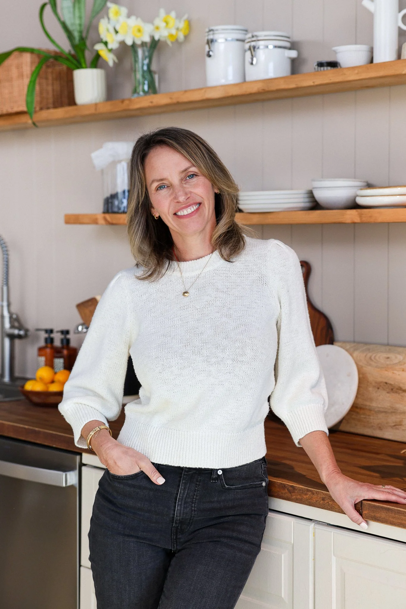 A woman with shoulder-length light brown hair, smiling, wearing a beige striped blouse, sitting in a kitchen or craft room with shelves and artwork in the background.