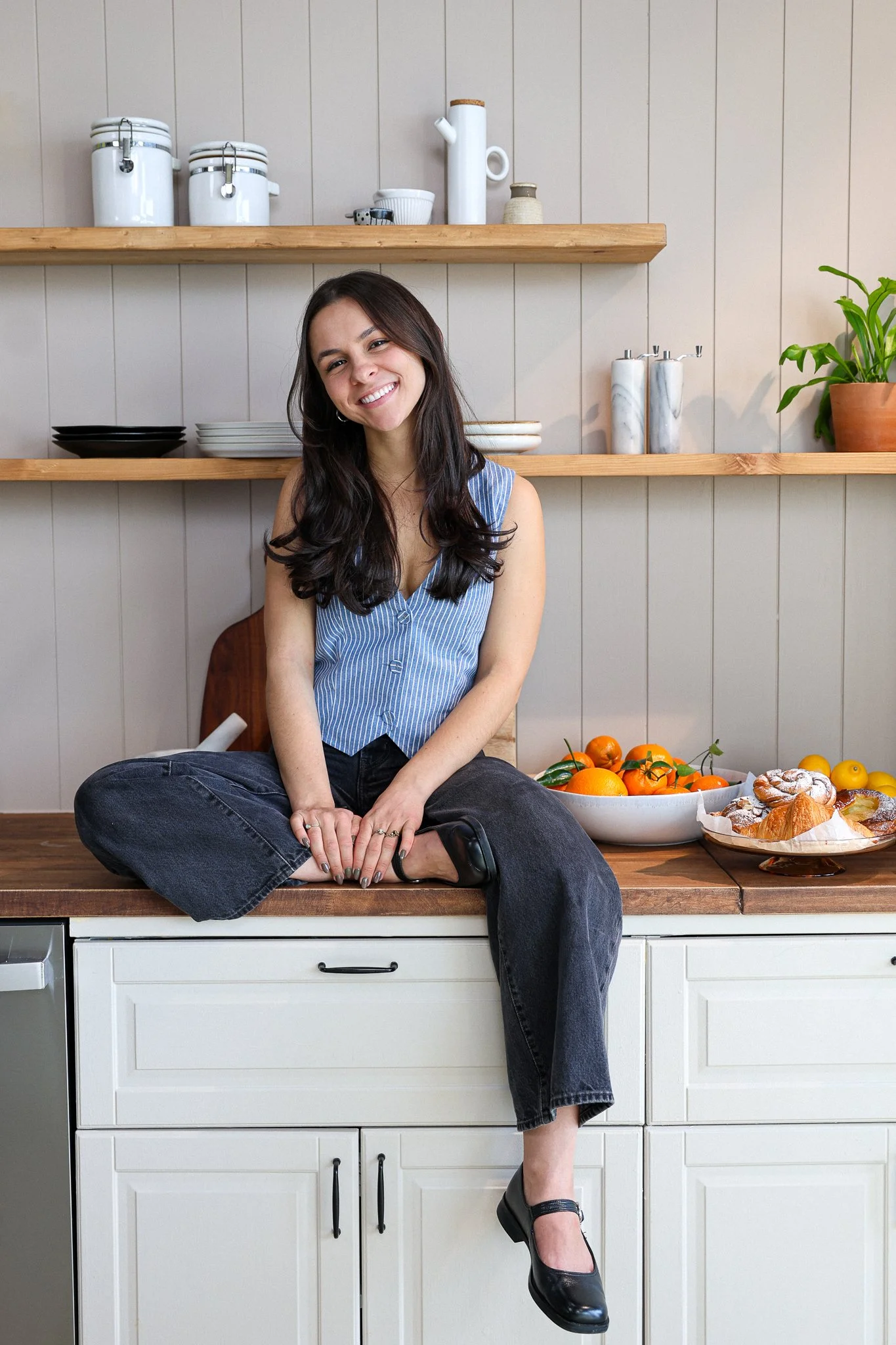 A young woman with dark hair pulled back, wearing a beige jacket with a black collar and a light turtleneck, smiling inside a cozy kitchen with shelves holding white bowls, a green plant, and a colorful tiled wall.