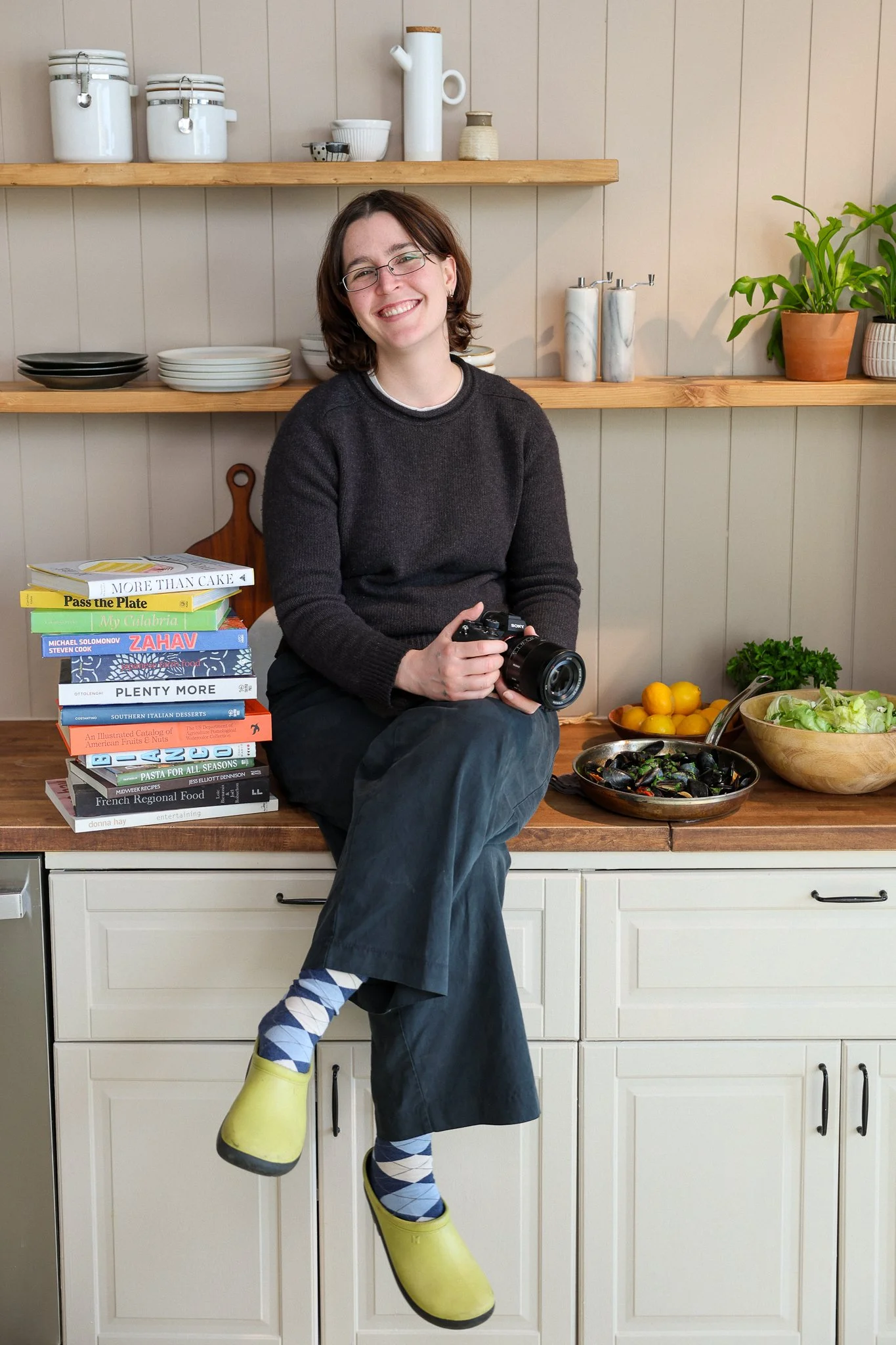 A woman with shoulder-length dark hair, glasses, and silver jewelry wearing a blue sweater, standing in a room with shelves holding kitchen cookware and decorative items.